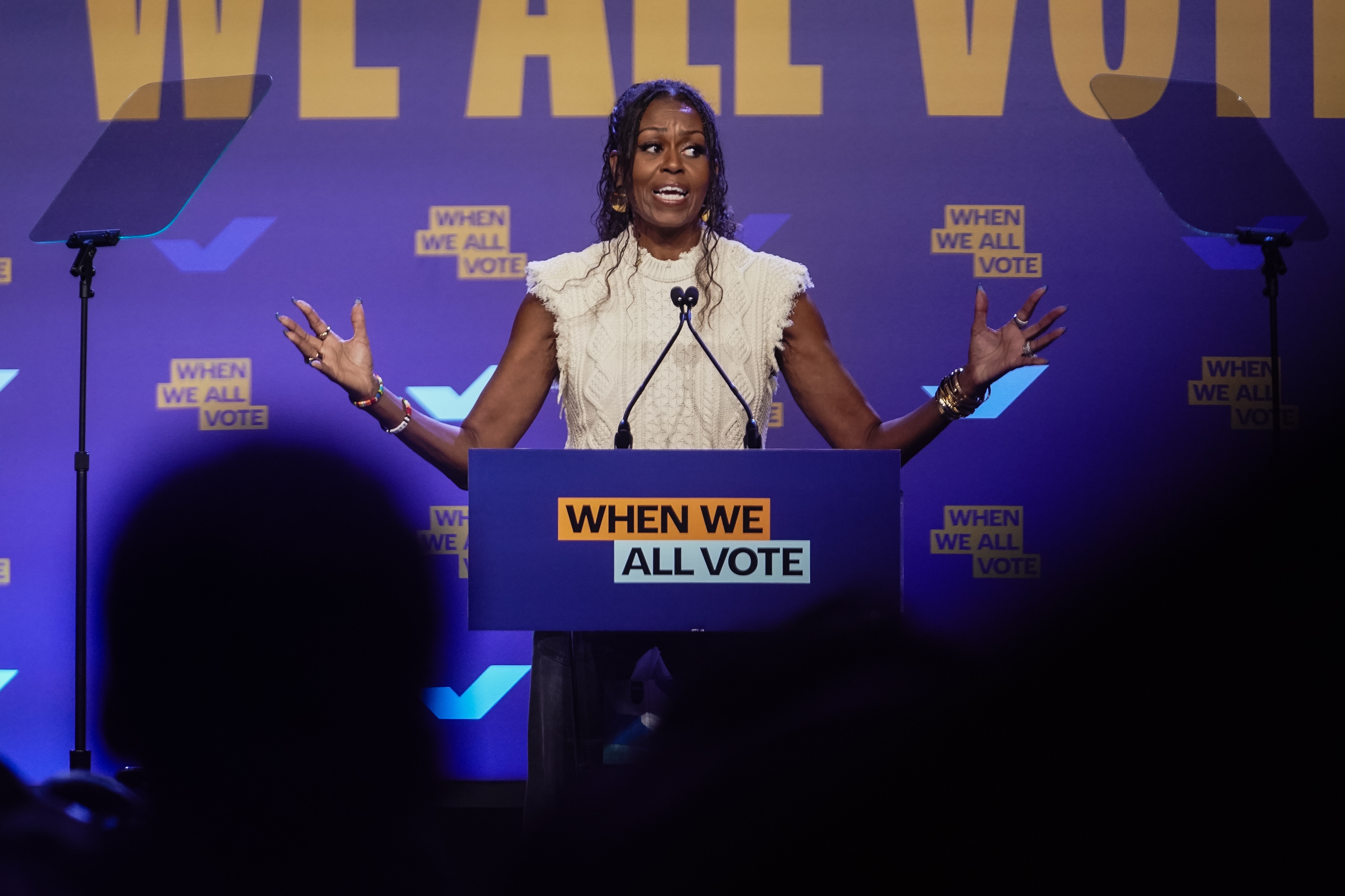 A person stands at a podium labeled "When We All Vote," speaking passionately with outstretched arms at an event