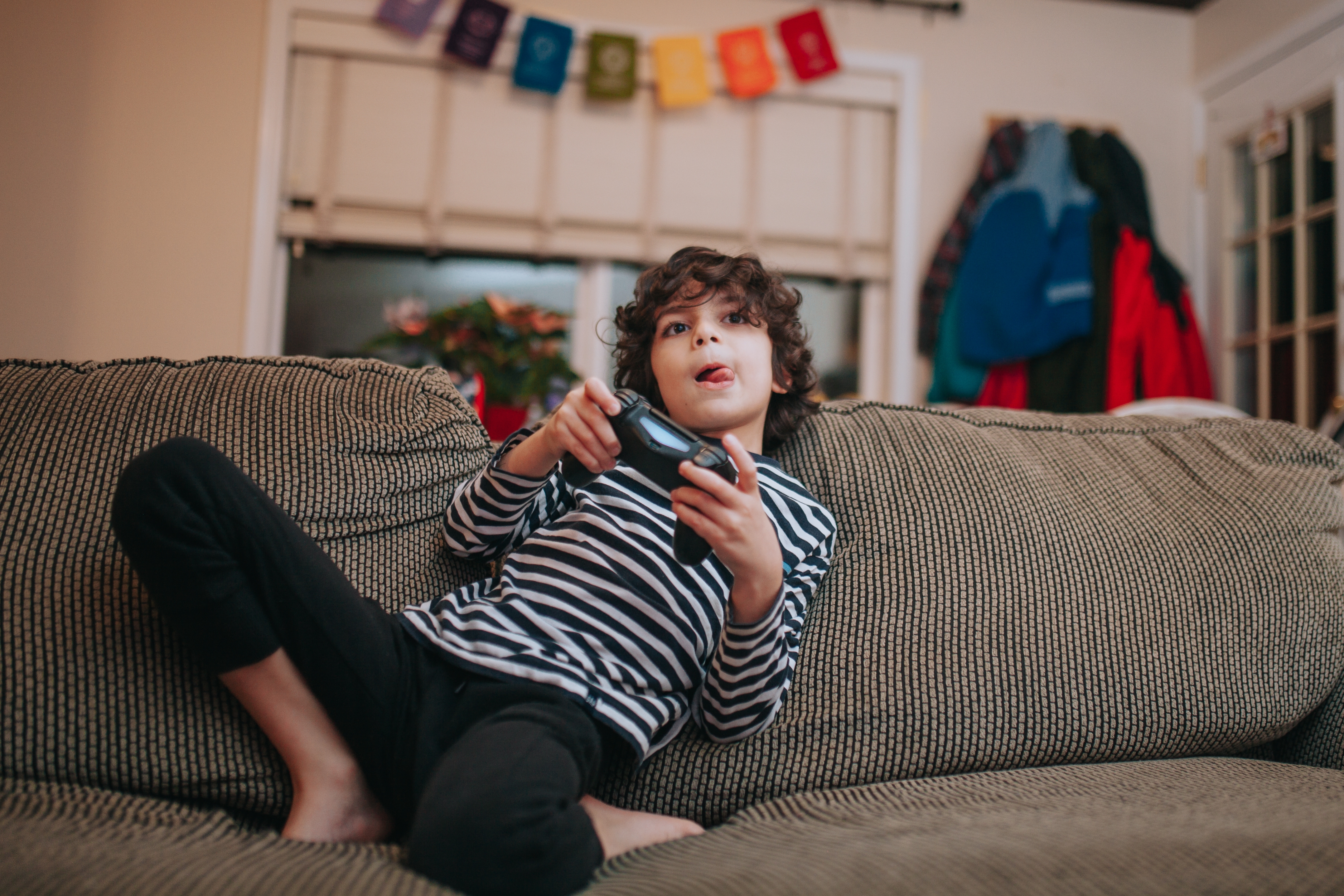 Child in a striped shirt playing video games on a couch, with colorful banner in the background