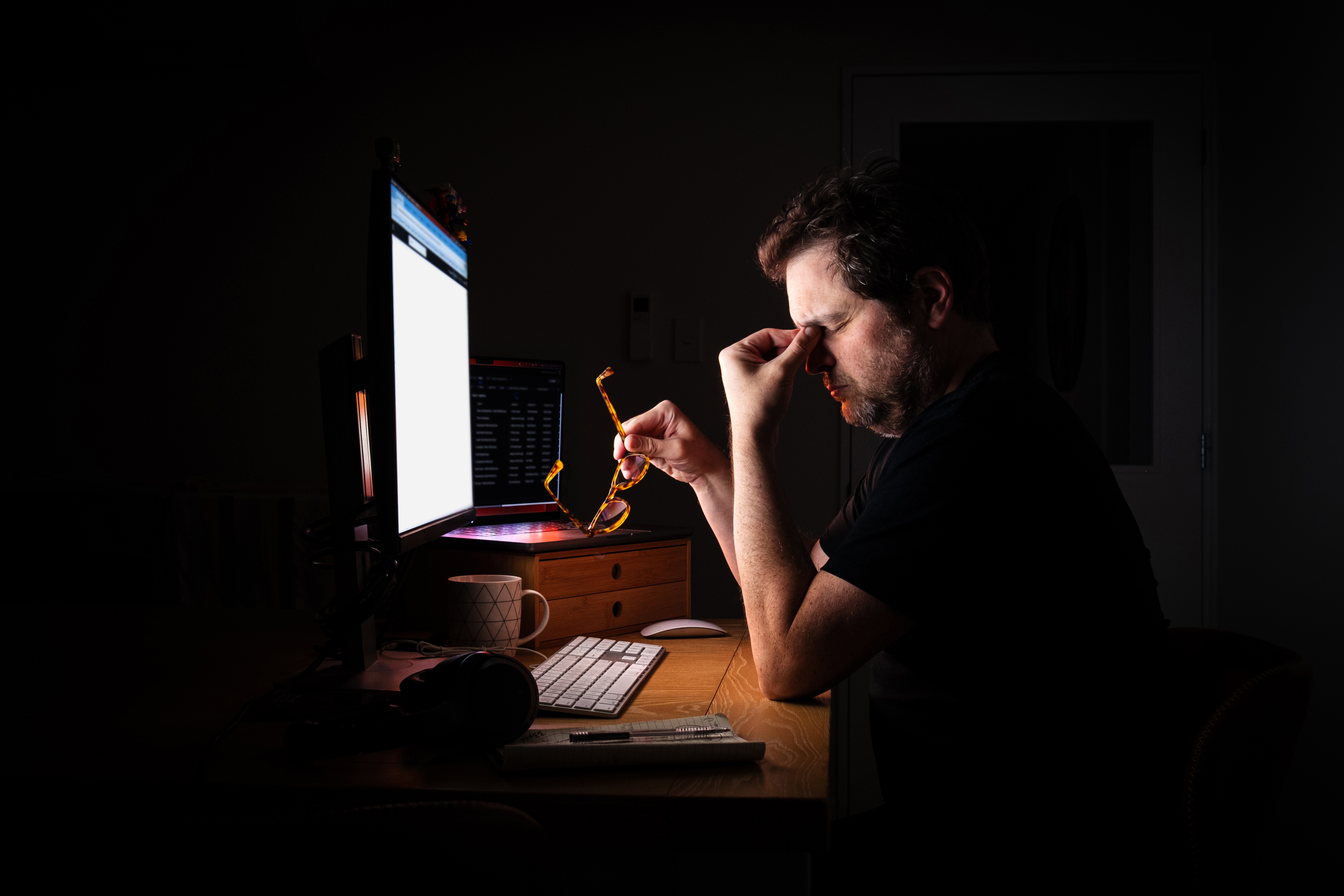 Person at a desk, holding glasses and looking tired in front of computer screens, suggesting late-night work or fatigue