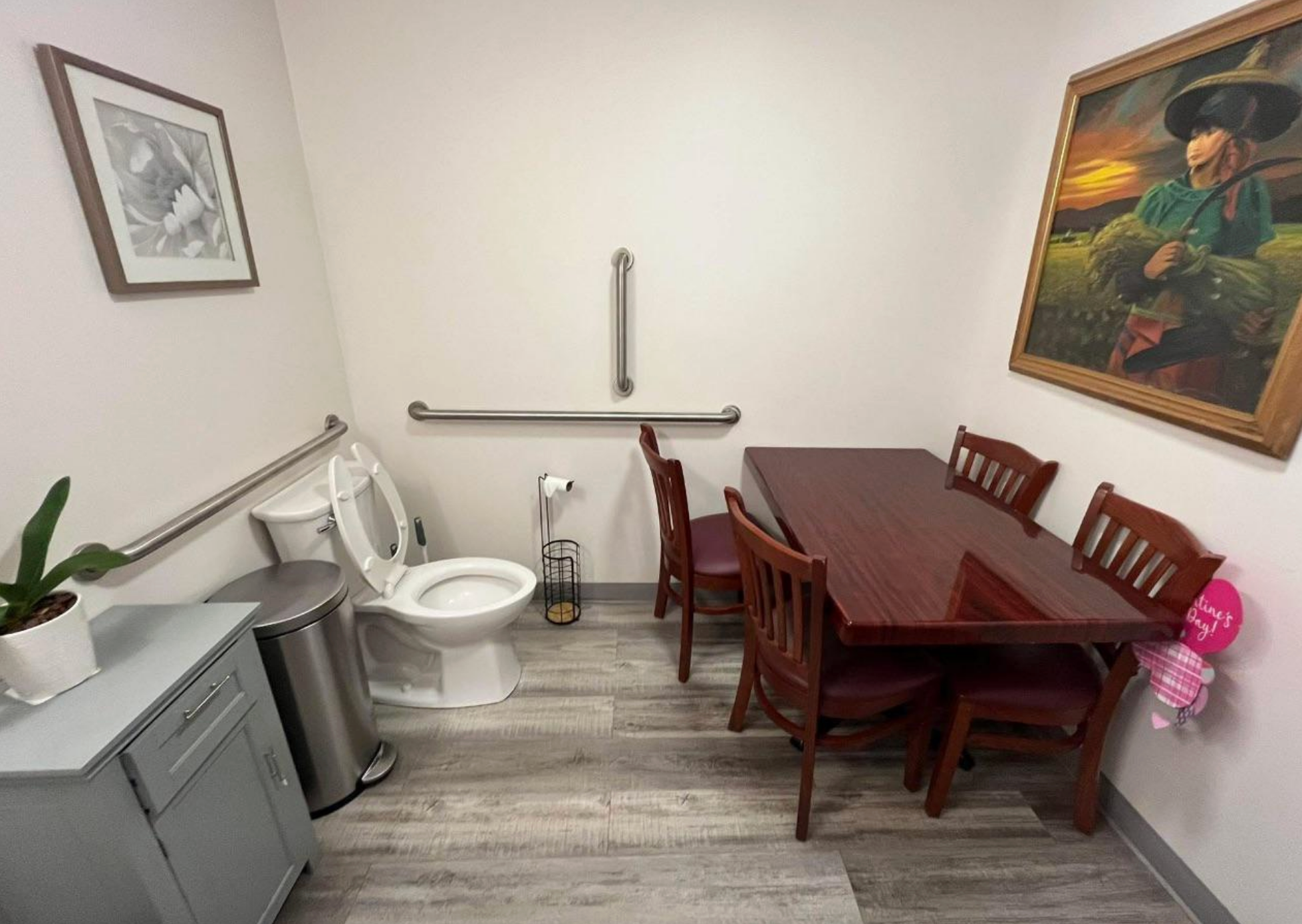 Room with a toilet, dining table, and chairs. Artwork and a plant decorate the walls. A mix of bathroom and dining area elements