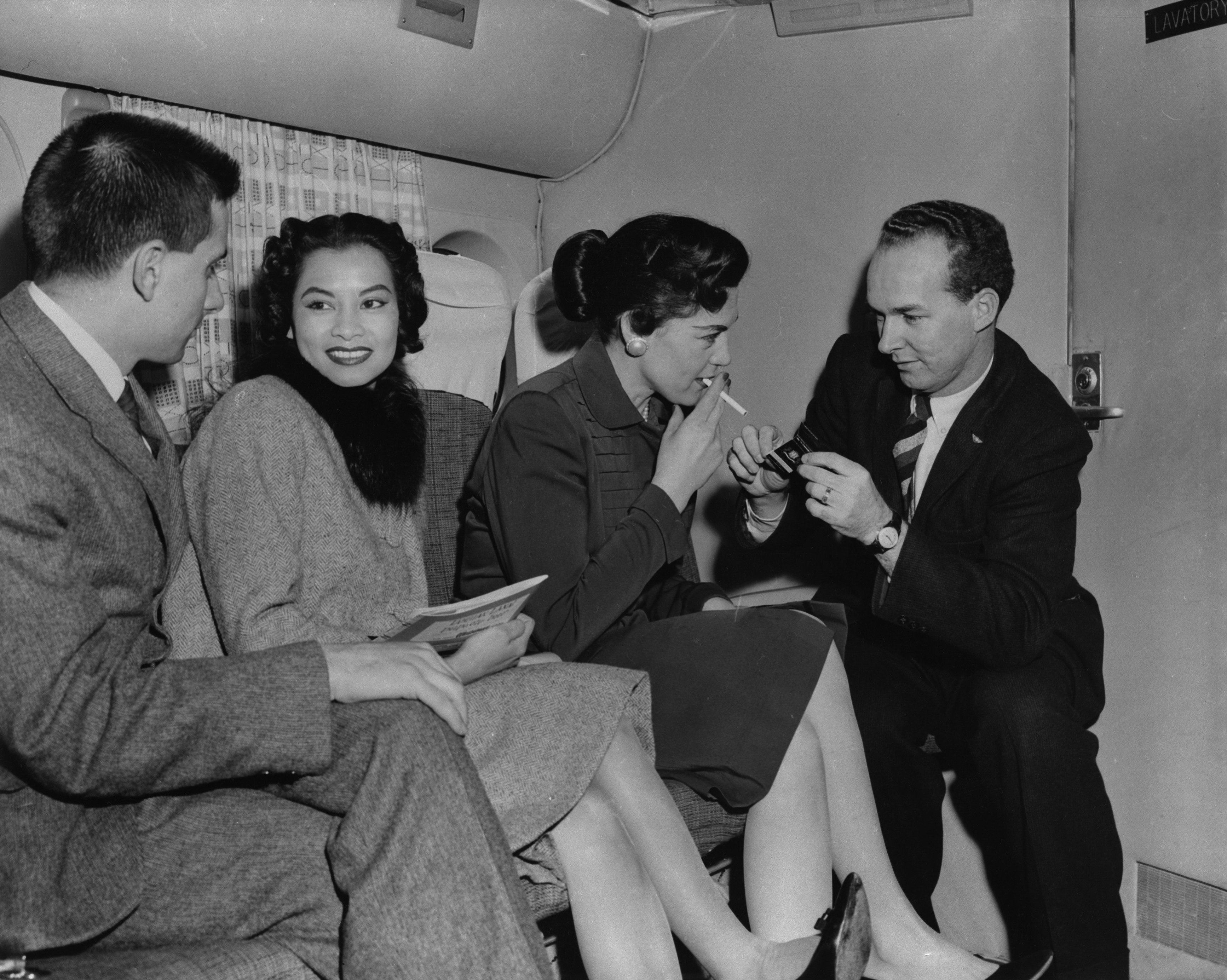 1950s airplane cabin scene: two women and a man in suits seated, a flight attendant lighting a cigarette for one woman