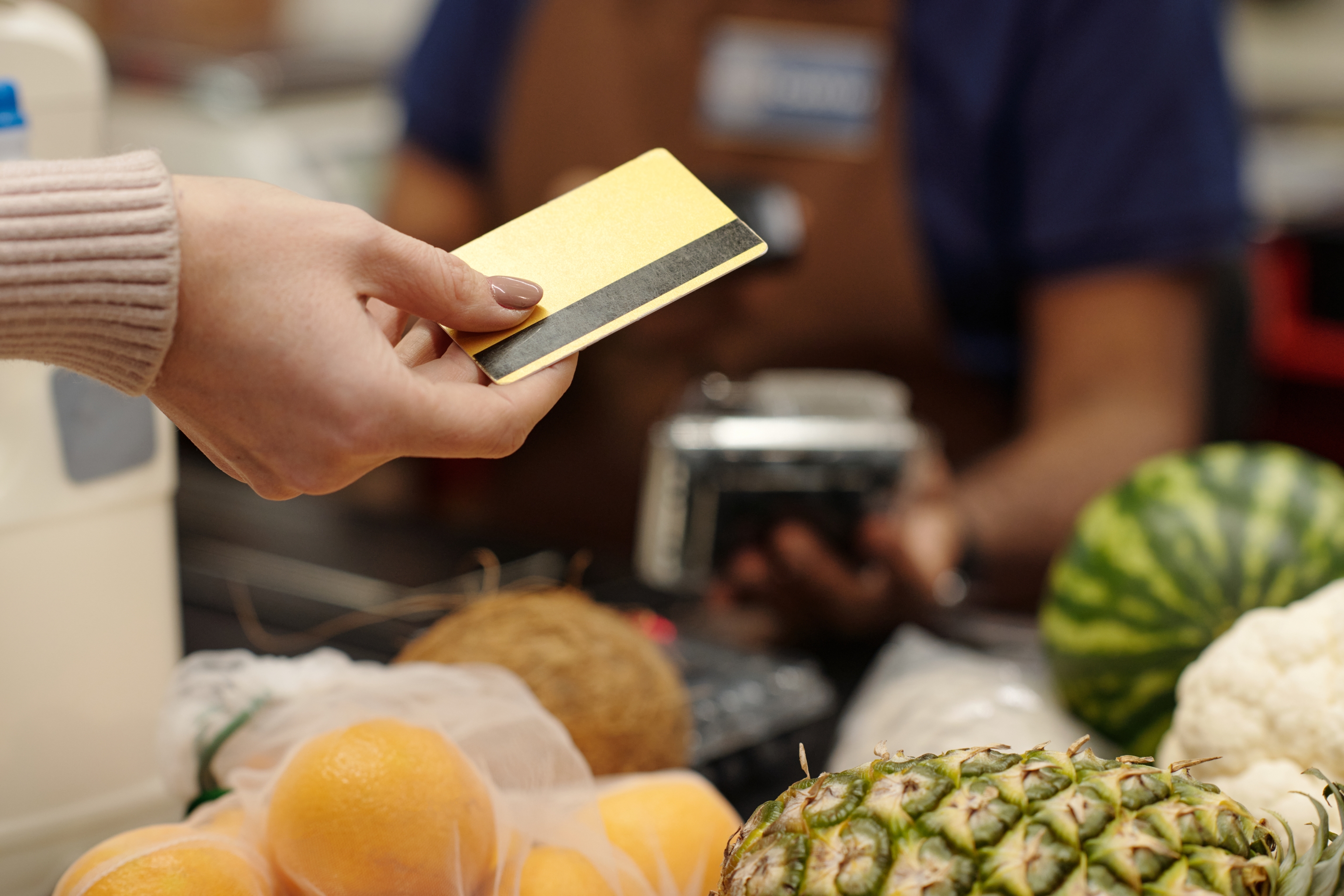 Person pays with a card at a grocery checkout, near fruits and vegetables. Cashier operates card machine