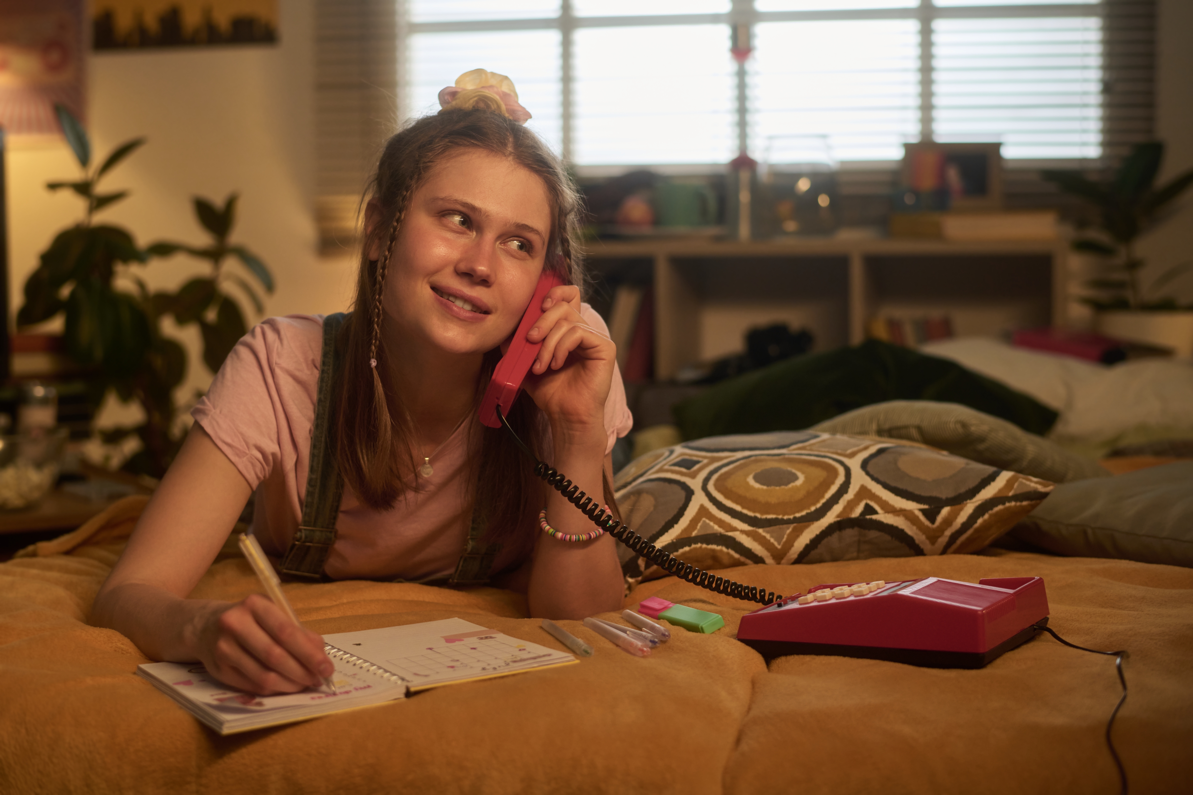 Person lying on bed, talking on a retro phone, and writing in a notebook, with a thoughtful expression. Cozy room background with plants and decor