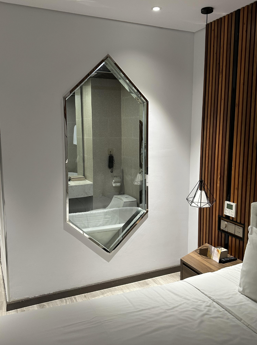 Modern hotel room with geometric mirror reflecting a bathroom. Features a wood-accented wall, a pendant lamp, and part of a bed. Minimalist decor