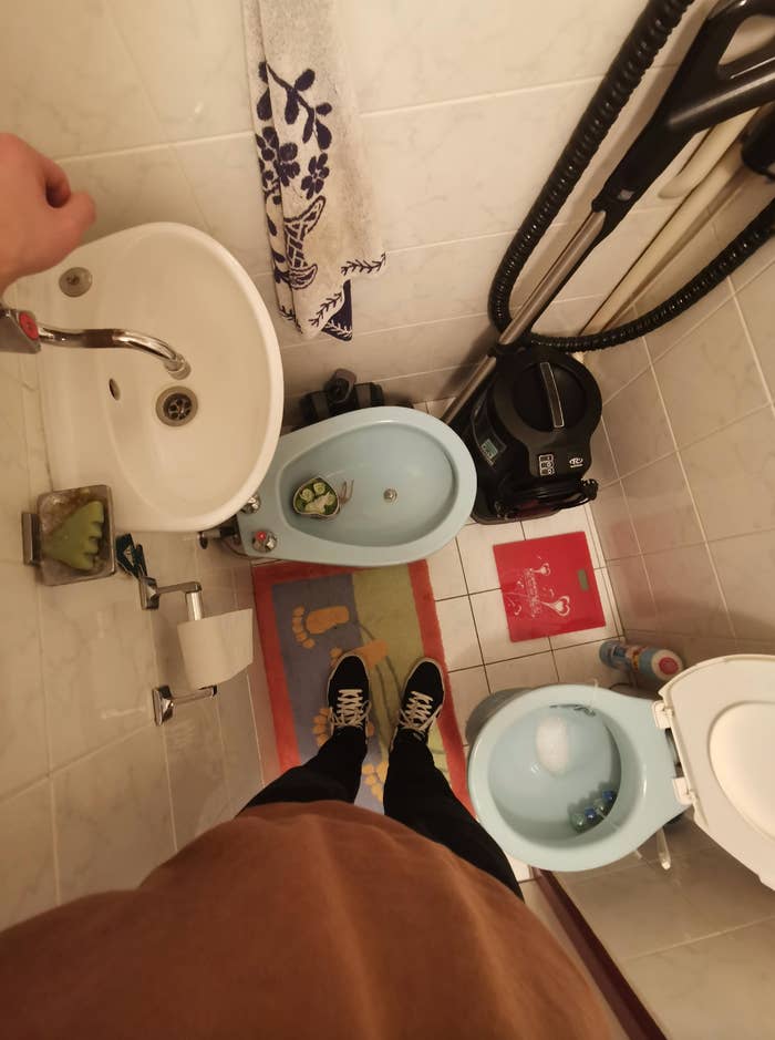 Bathroom with sink, bidet, vacuum cleaner, and toilet. Person's feet in sneakers visible