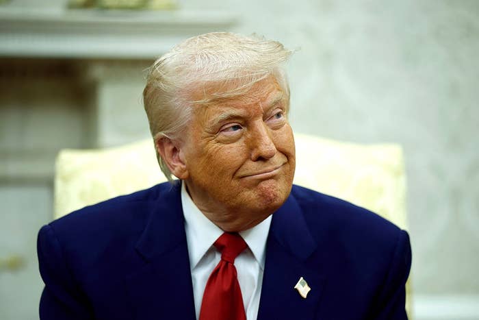 A person in a suit and red tie is sitting indoors, looking to the side with a slight smile