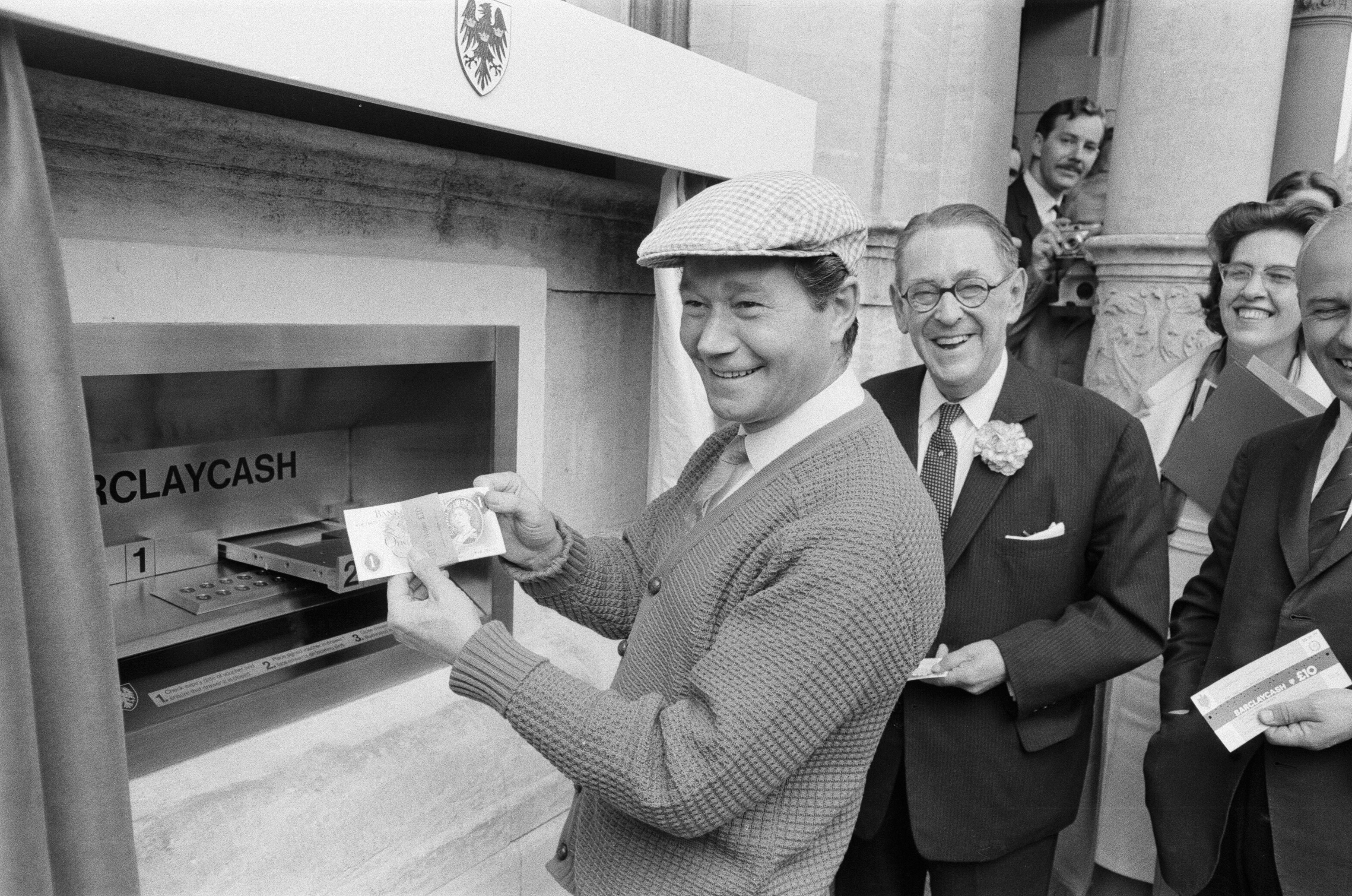 Man in flat cap withdraws cash from an early ATM, surrounded by smiling onlookers in suits, showcasing a historical banking moment