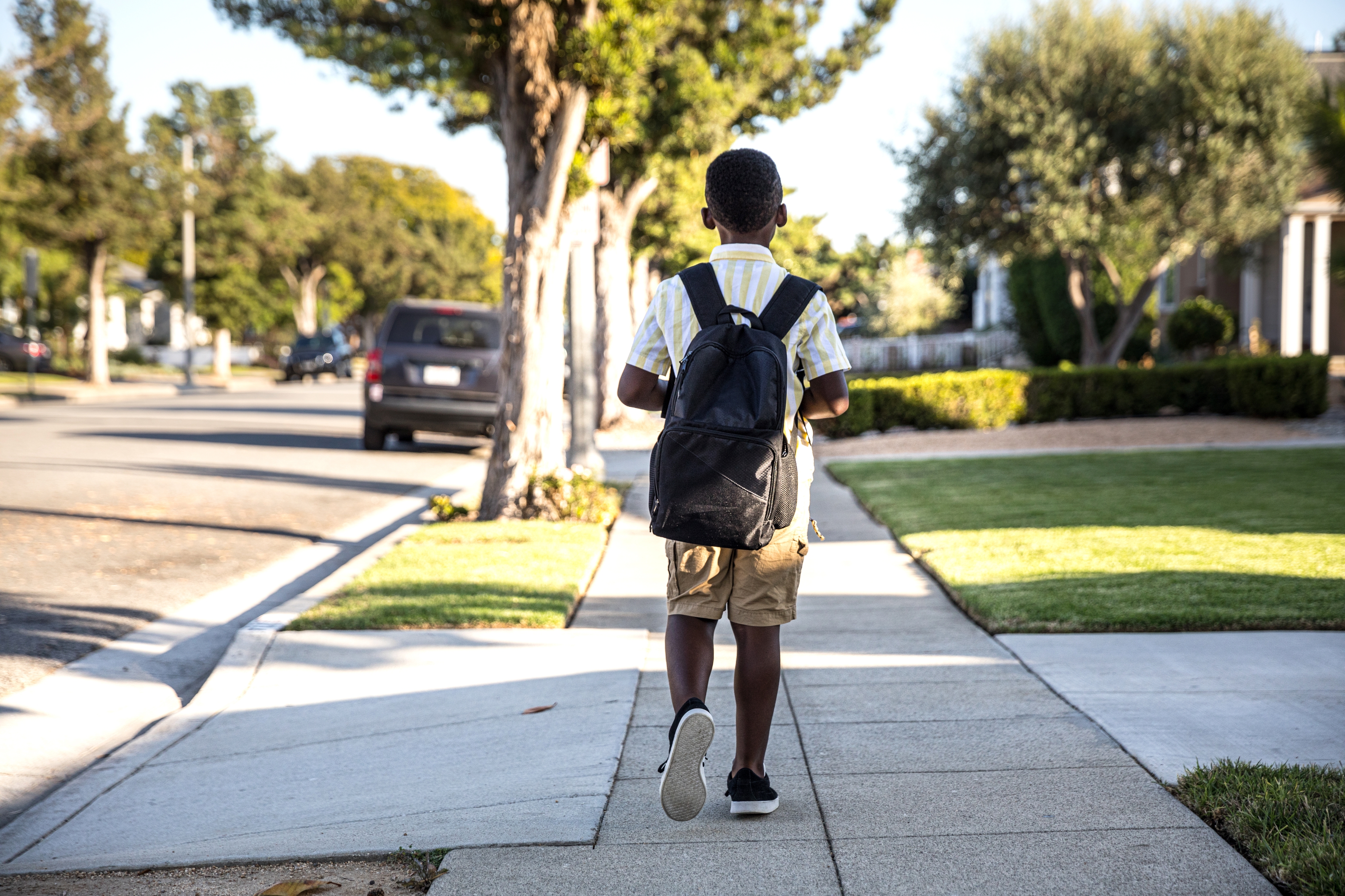 Child walking on a suburban sidewalk with a backpack, heading towards school. Trees and parked cars line the street