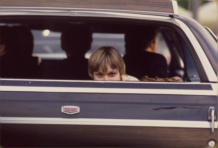 A person peeks through the window of a car, with others sitting inside, creating an air of curiosity and secrecy
