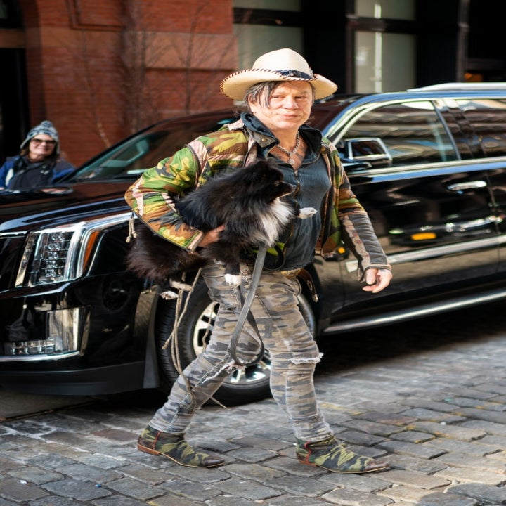 Person in a camouflage jacket and skinny jeans walking a fluffy dog on cobblestone street. Stylish wear includes a wide-brimmed hat and loafers