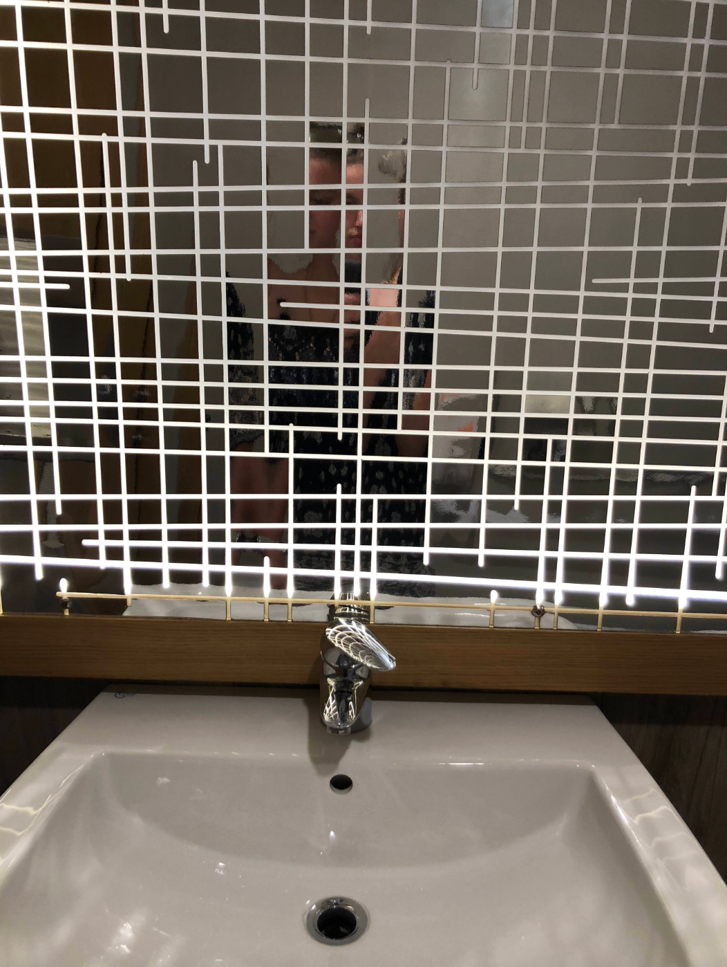 Person taking a selfie in front of a decorative mirror above a sink. The mirror features a geometric pattern