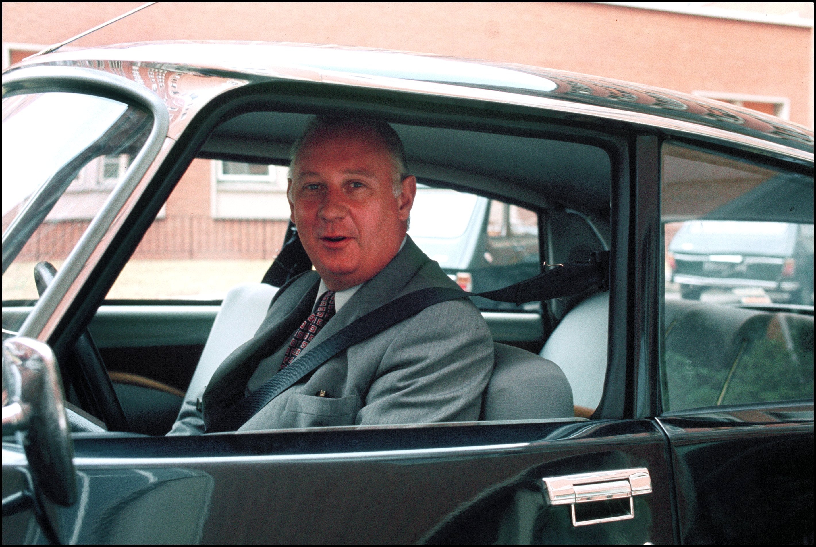 A person in a suit sits in a vintage car, looking out the window, with a classic building in the background