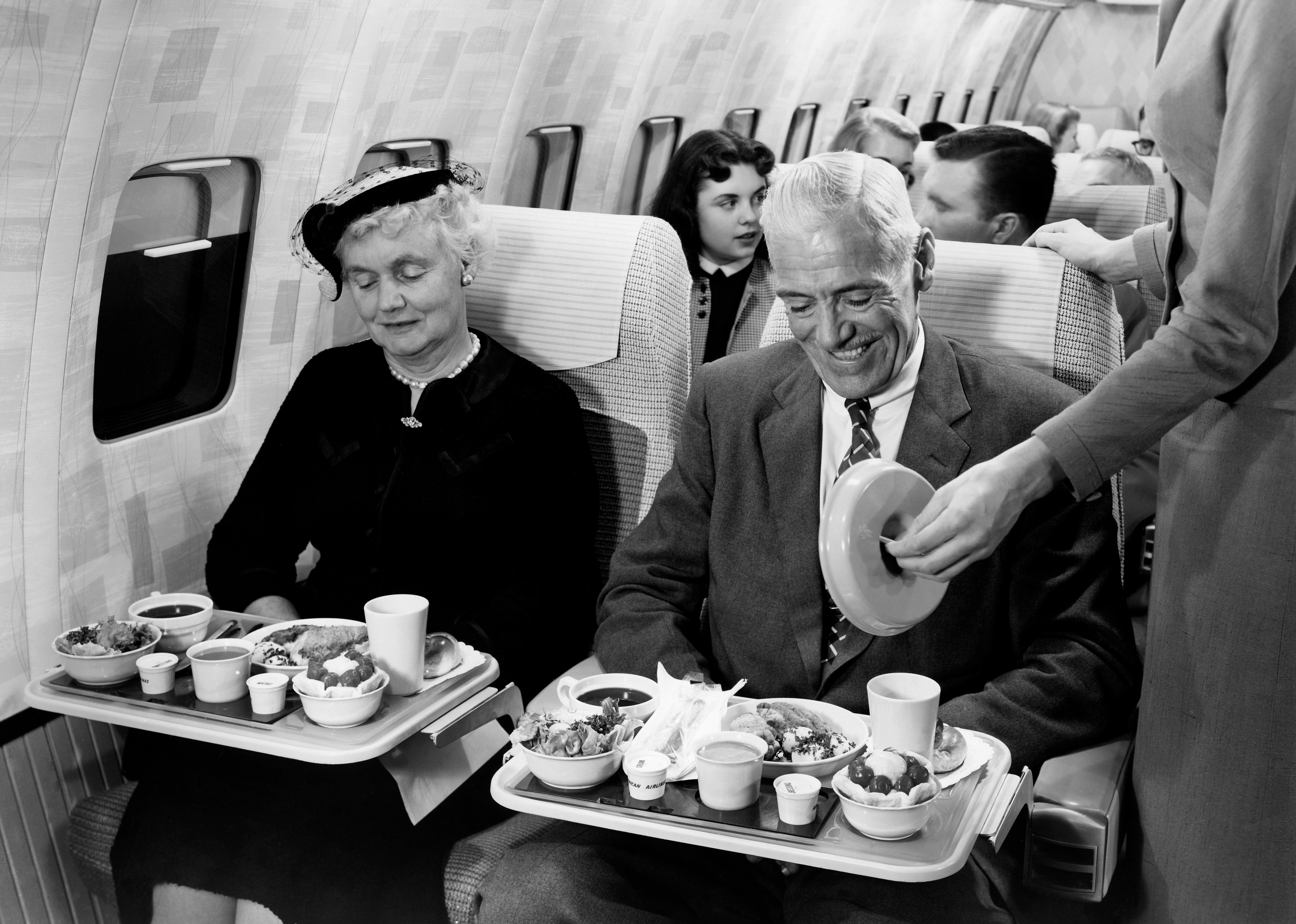 Passengers in a mid-20th century airplane enjoy a meal service. A flight attendant assists at one passenger's tray