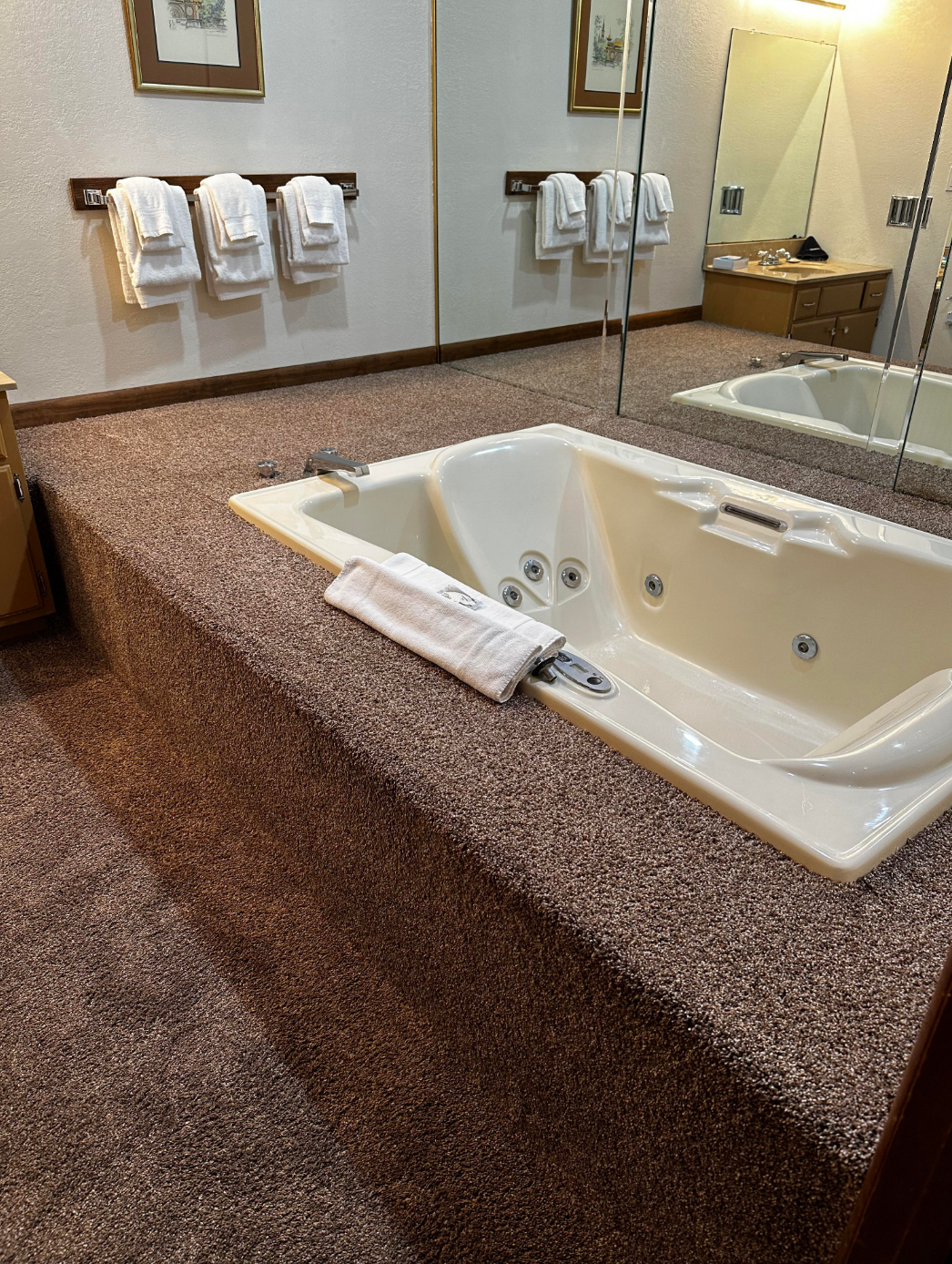 A mirrored bathroom features a sunken Jacuzzi tub on a carpeted platform, with neatly folded towels on racks and a wooden vanity nearby
