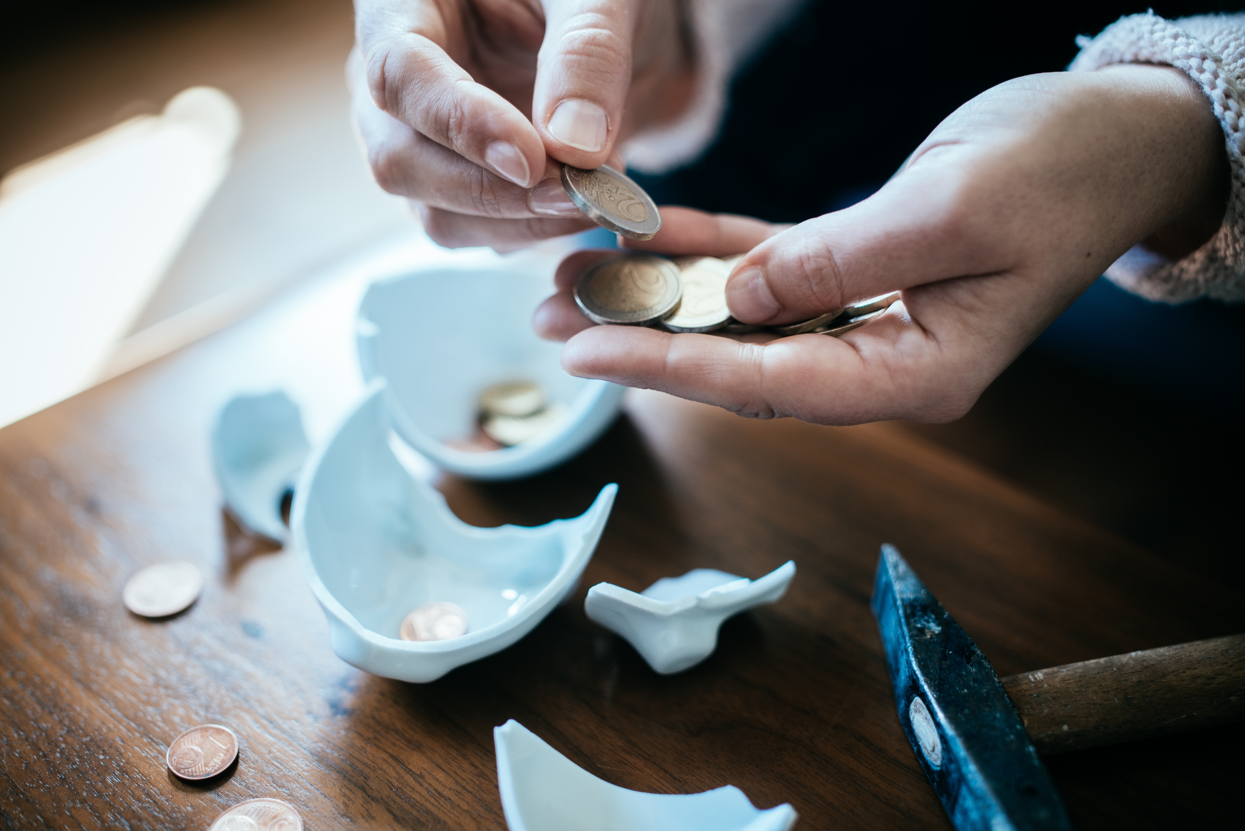 Person counting coins from broken piggy bank on a wooden table with a hammer nearby
