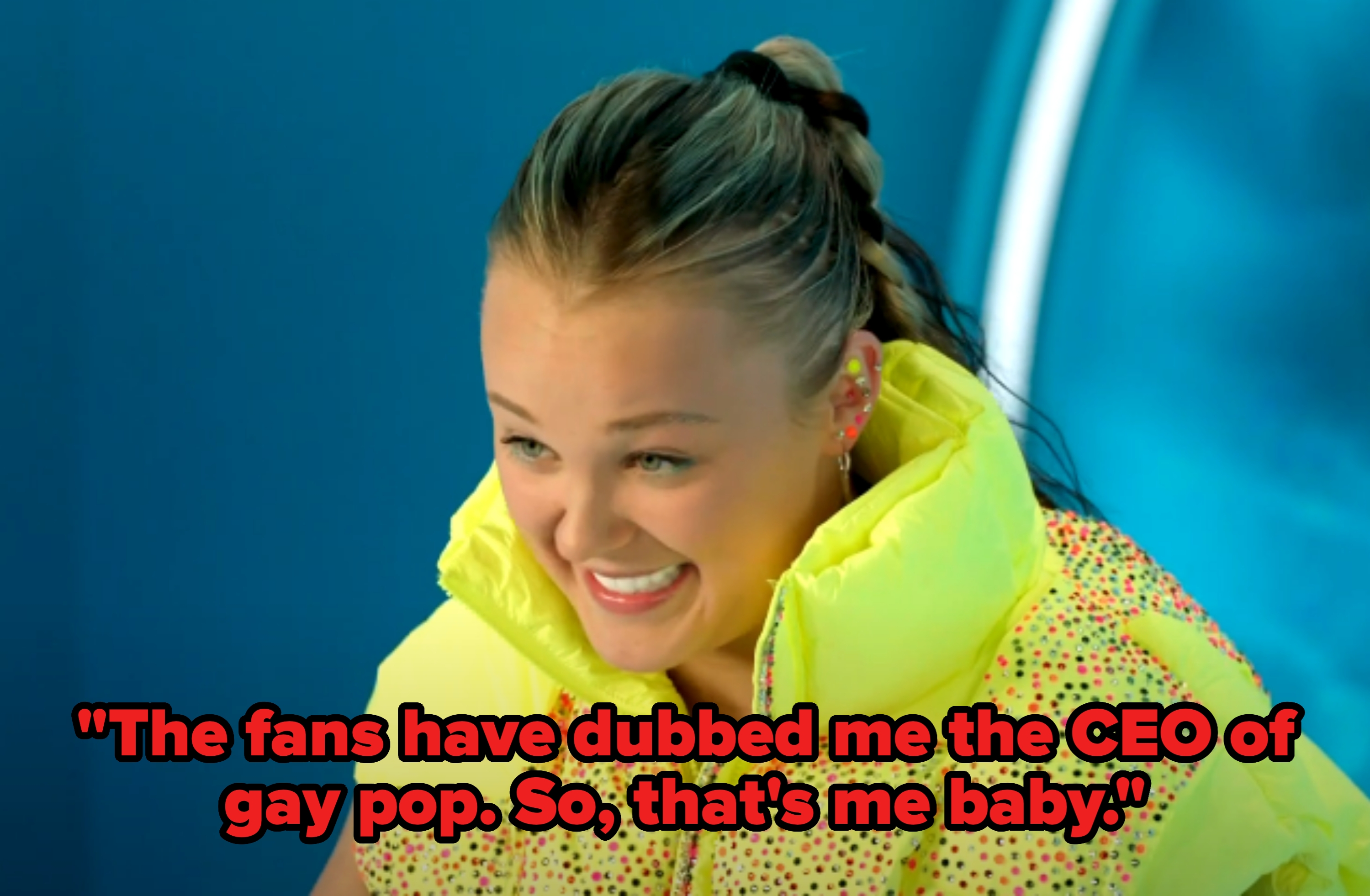 Person smiling, wearing a bright, puffy vest with a pattern. Hair is tied back, setting is indoors with a blurred background