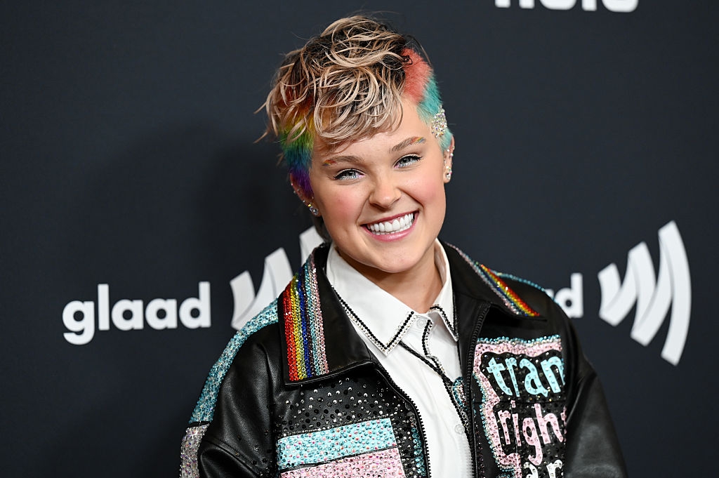 Person with short, styled hair and colorful accents smiles on the red carpet, wearing a studded jacket with text and sparkling details