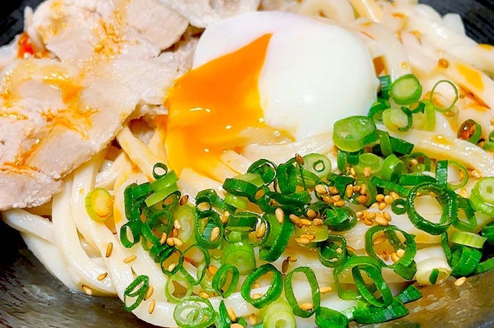 うどんの上にとろけた卵、薄切りの肉、刻んだネギ、ゴマがのっている料理。
