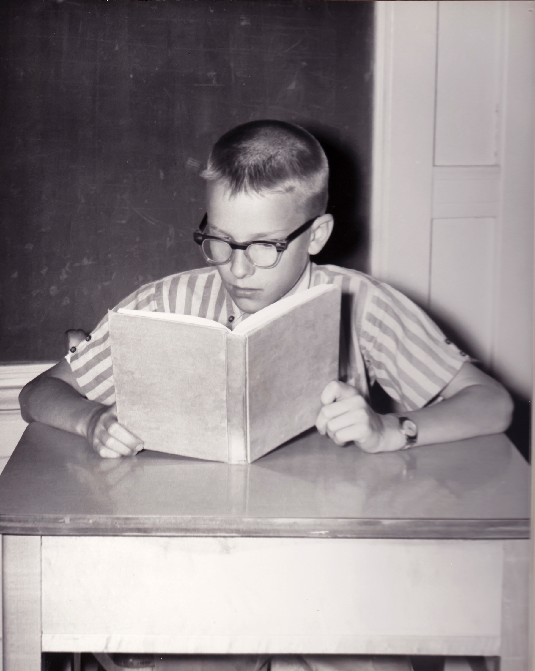 A young boy with glasses sits at a desk, intently reading a book in a classroom setting