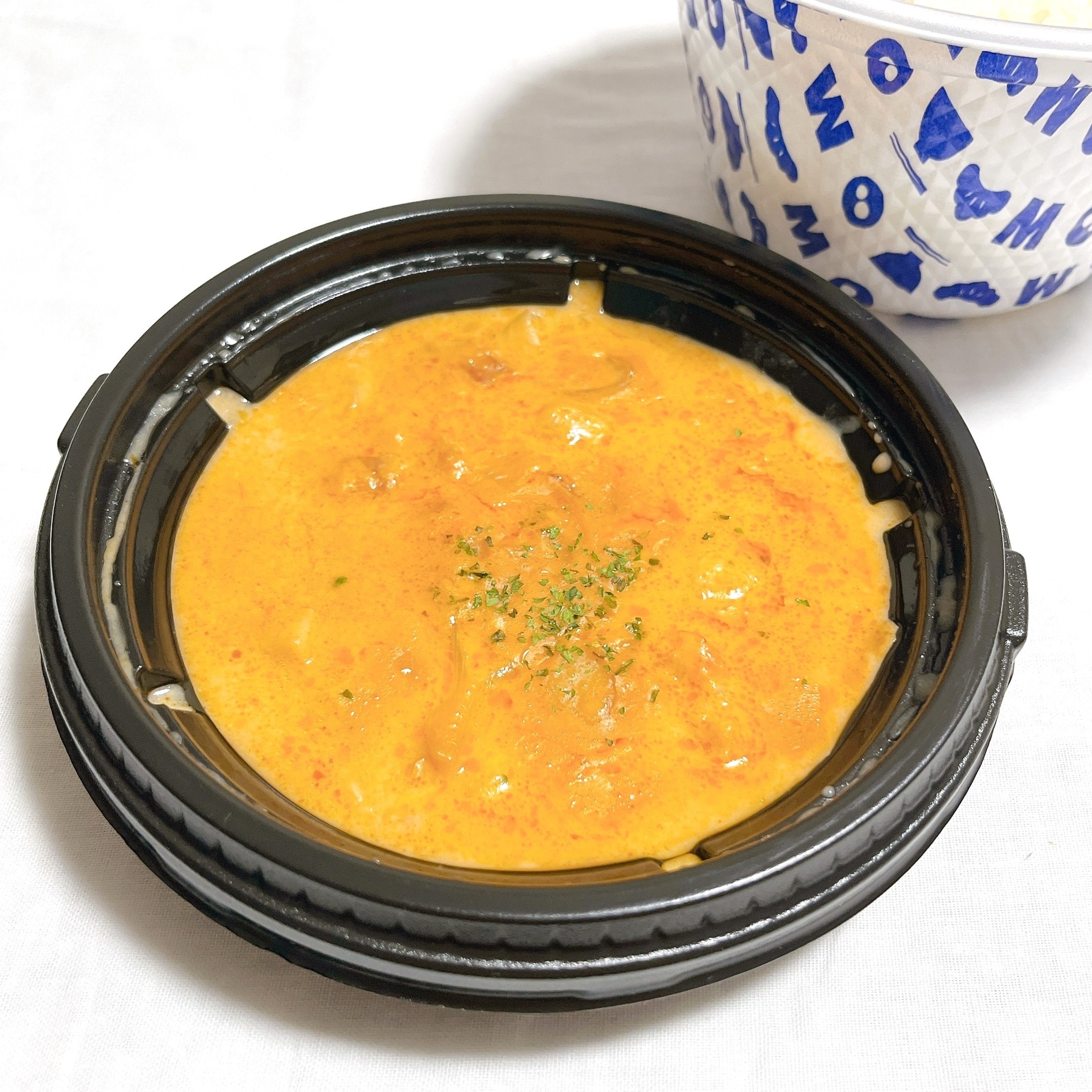 クリーミーなカレーが黒い容器に入っている様子。隣には青い模様が入った白いボウル。