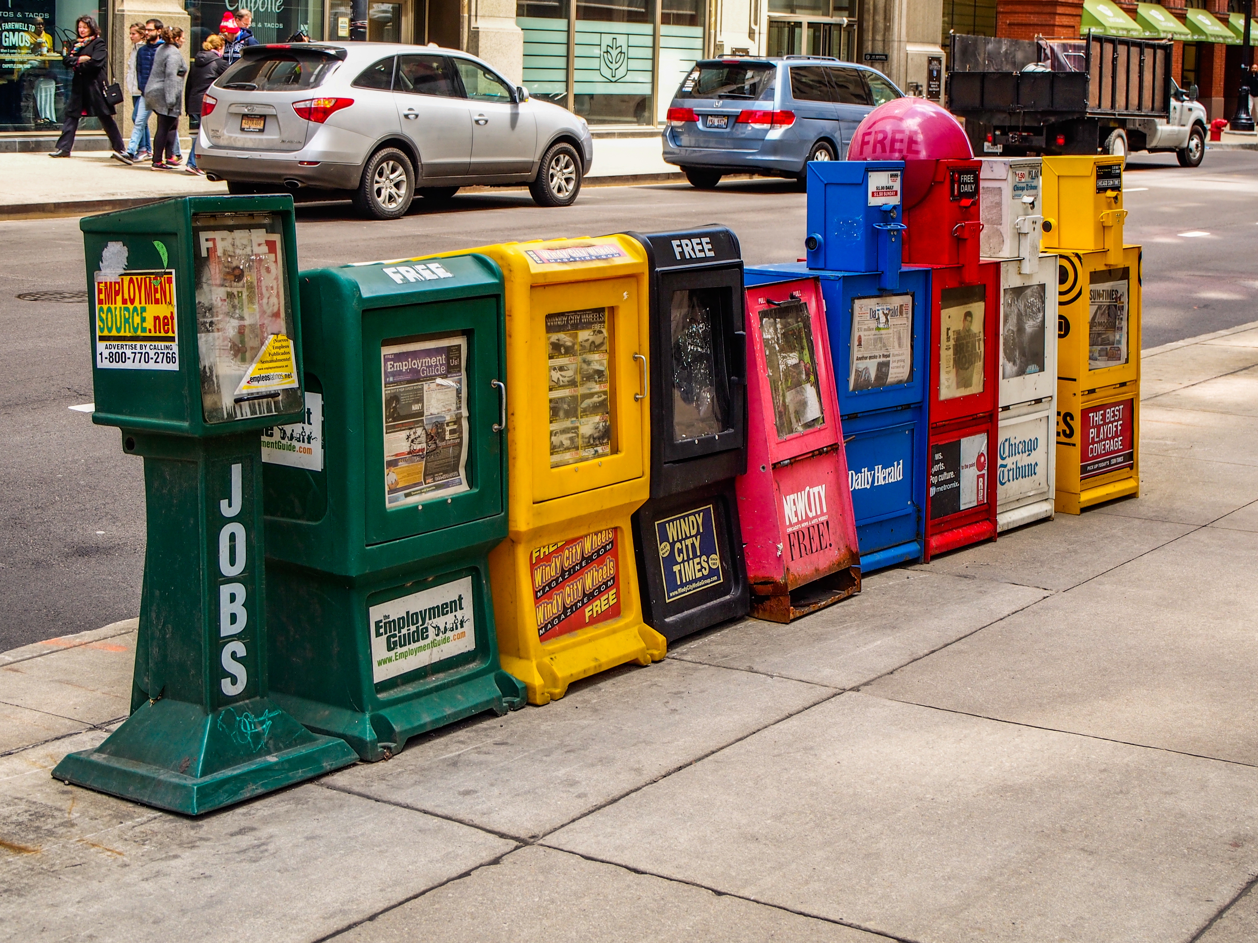 A row of colorful newspaper vending machines on a city sidewalk, offering various local and national publications. Cars and storefronts are in the background