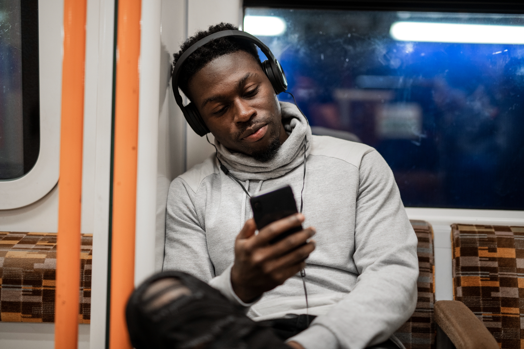 A man in a hoodie listens to music on headphones, sitting in a train with a phone in hand, appearing relaxed and absorbed