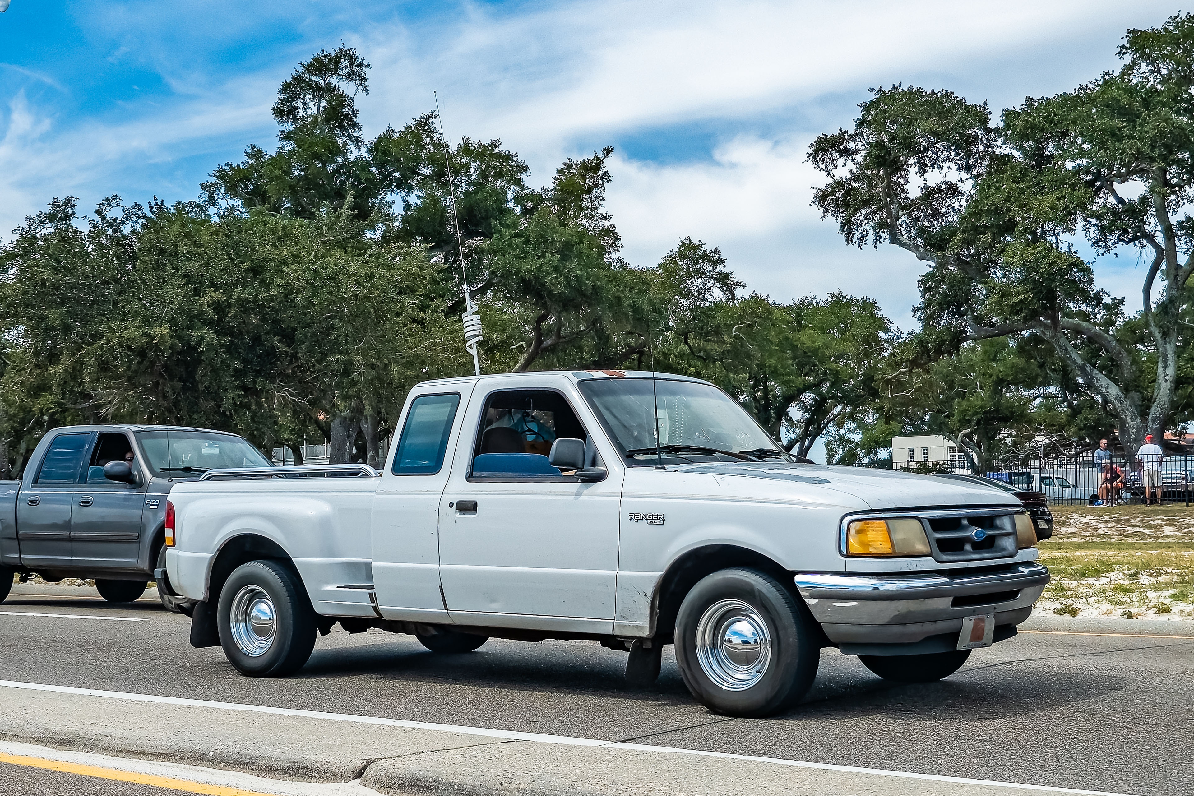 Two pickup trucks driving on a road beside trees, one white and one gray, with a partly cloudy sky in the background
