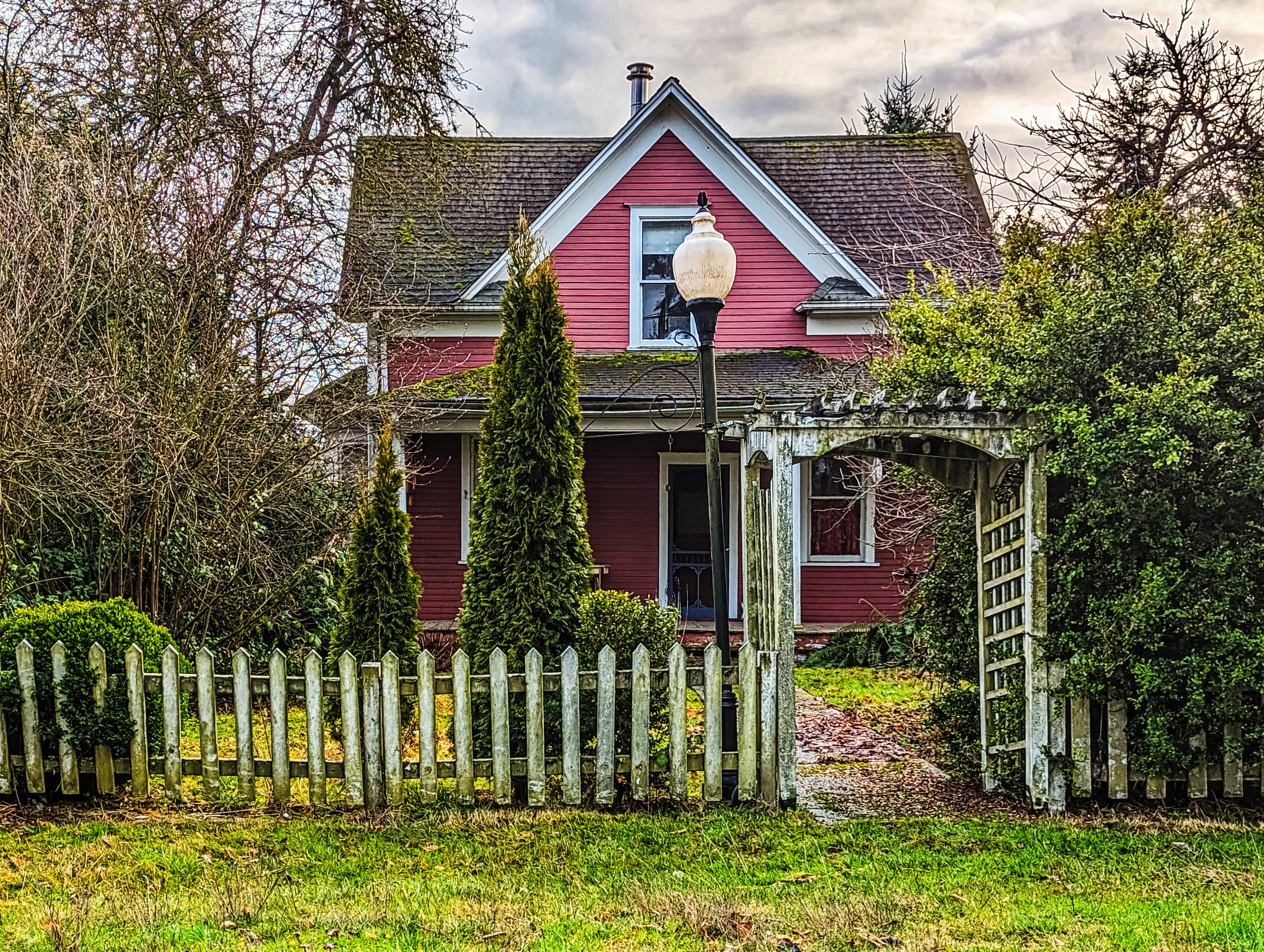A quaint house with a porch, a pitched roof, and a white fence gate surrounded by trees and bushes. A vintage-style streetlamp stands in front