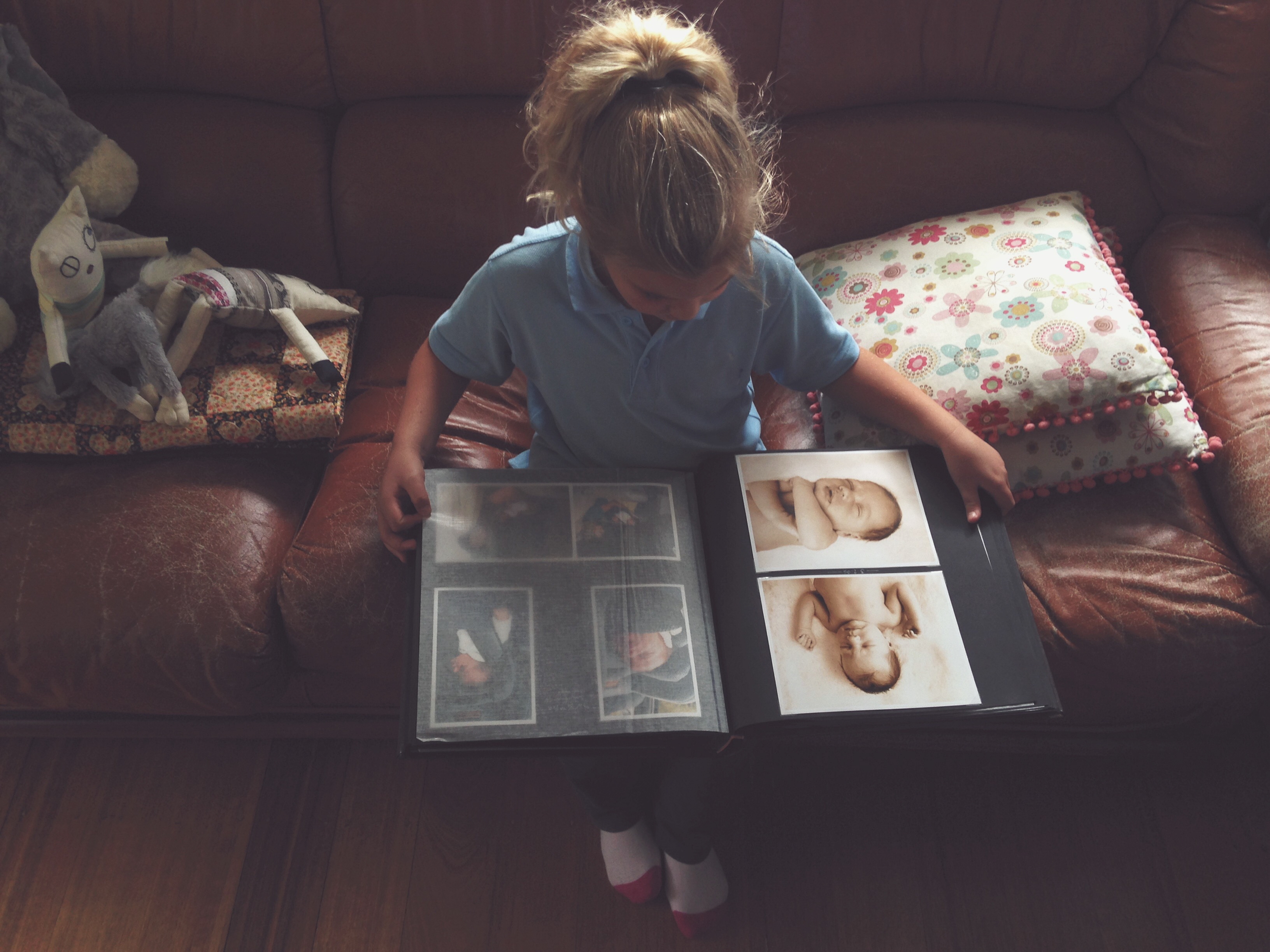 Child sitting on a couch, looking at a photo album containing baby pictures. Toys are on the couch beside them