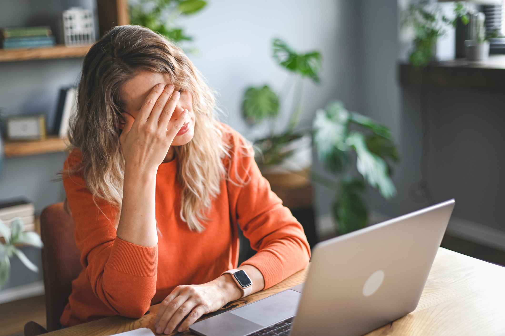 Person sitting at a desk with a laptop, hand on forehead, looking stressed or frustrated. Indoor plants in the background