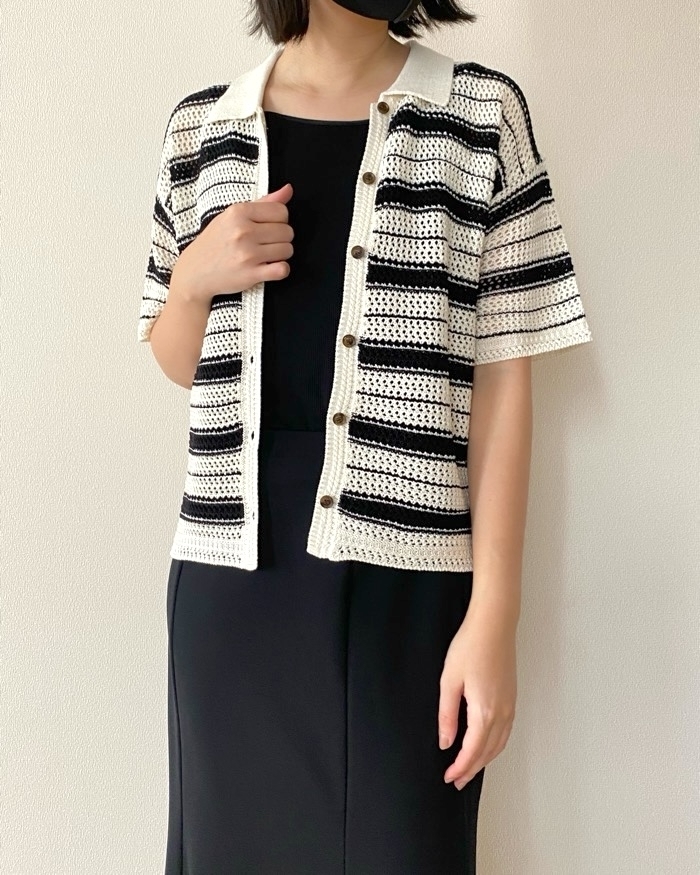 黒いワンピースの上にストライプのカーディガンを着た人物が立っている。