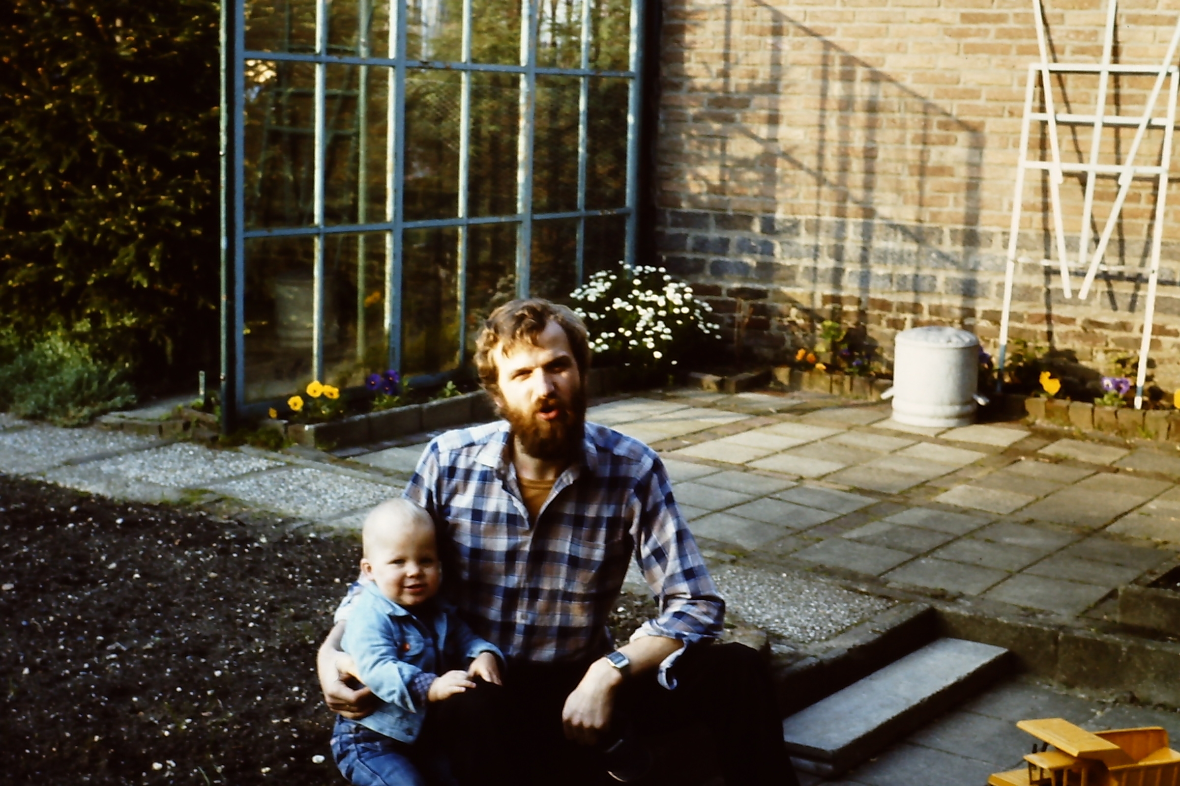 A man with a beard sits on steps holding a baby in an outdoor area with plants and a brick wall