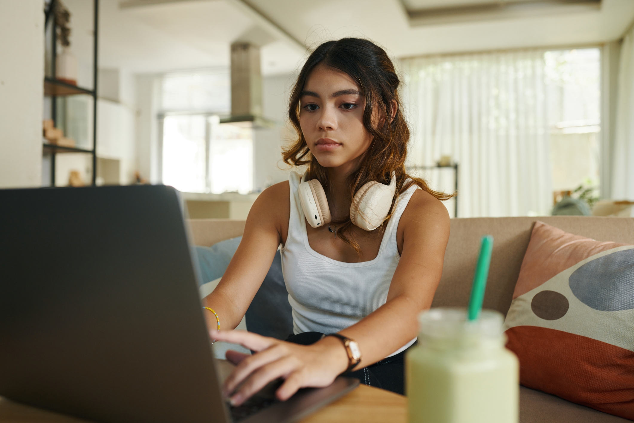 A person wearing headphones, seated in a living room, working on a laptop with a beverage nearby on the table