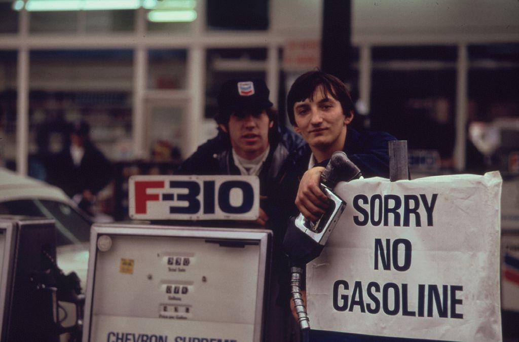 Two gas station attendants lean on a pump with a sign reading &quot;Sorry No Gasoline&quot; during a fuel shortage event