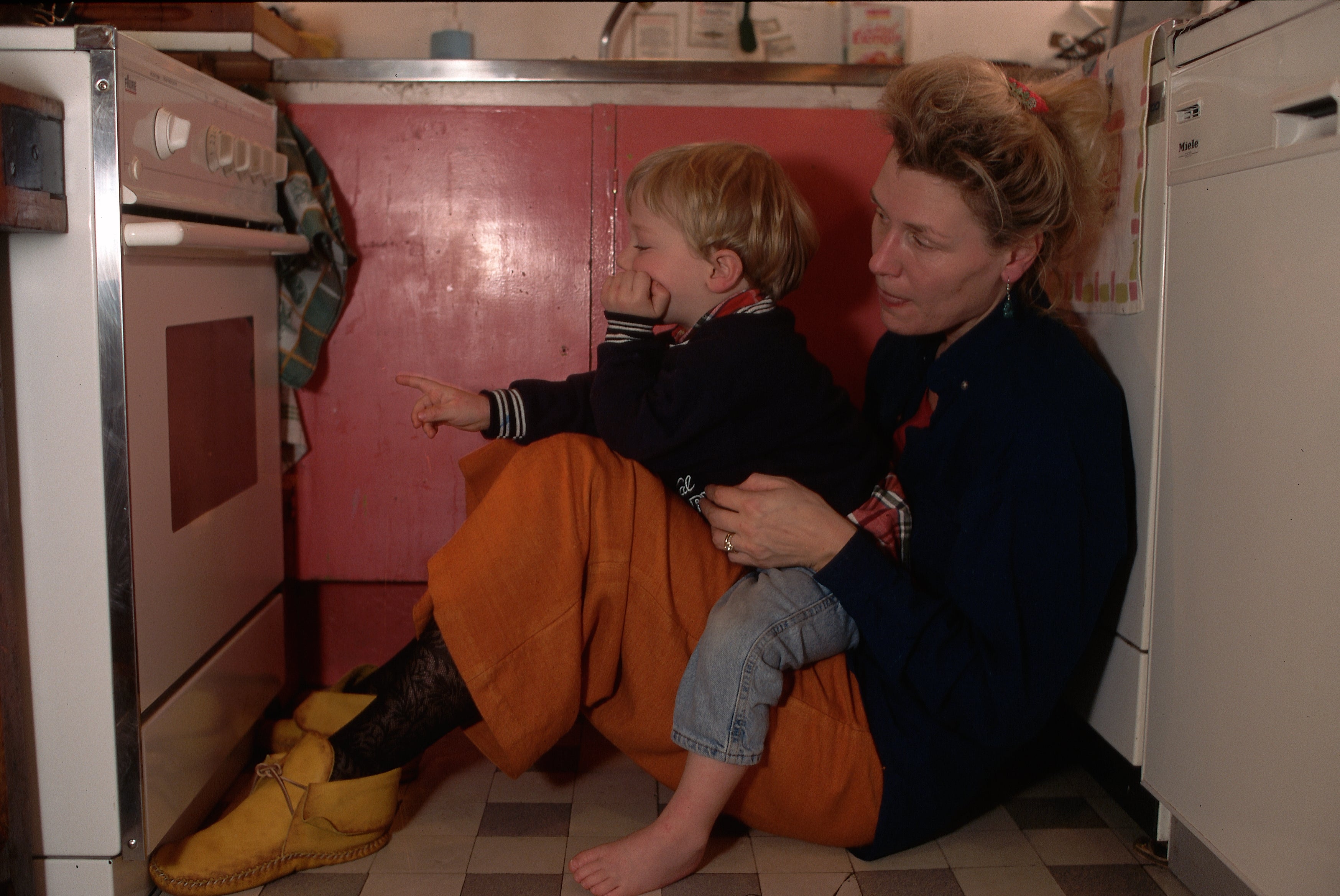 A woman sits on a kitchen floor with a child on her lap, both looking at an oven. The child points at the appliance. Casual, home setting