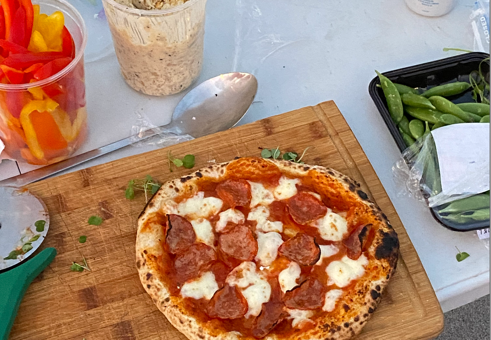 Pizza with pepperoni and cheese on a wooden board, surrounded by sliced peppers, dip, and snap peas on a table