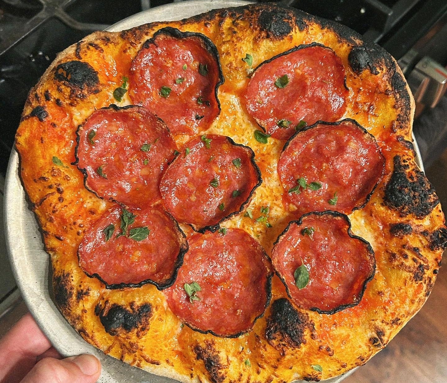 Hand holding a homemade pepperoni pizza with a crispy crust and salami