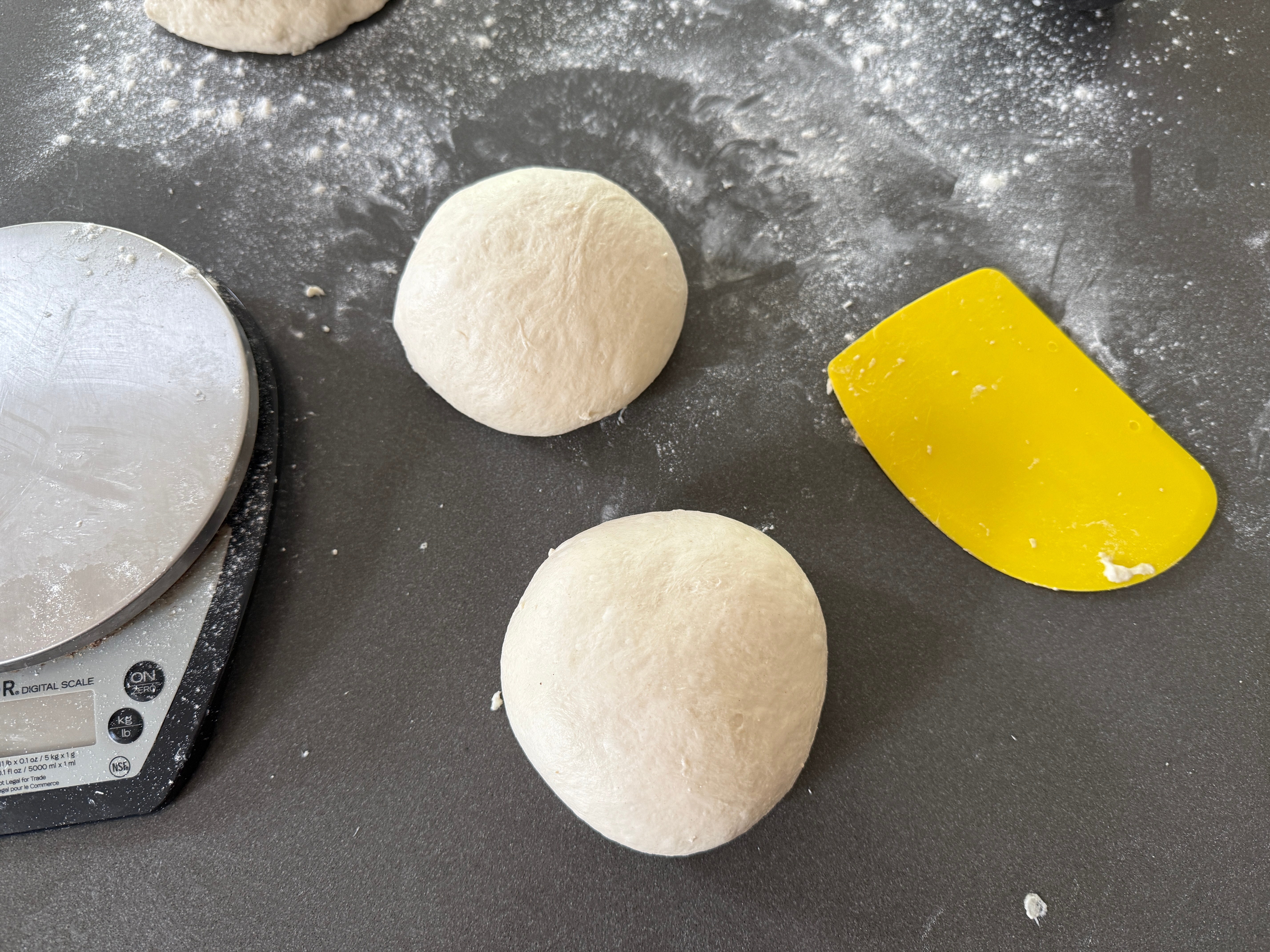 Dough balls on a floured counter beside a digital scale and a yellow dough scraper