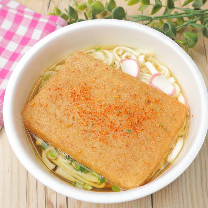 うどんの上にきつね揚げが乗った料理。ネギやかまぼこがトッピング。バックにピンクチェック柄の布と観葉植物。