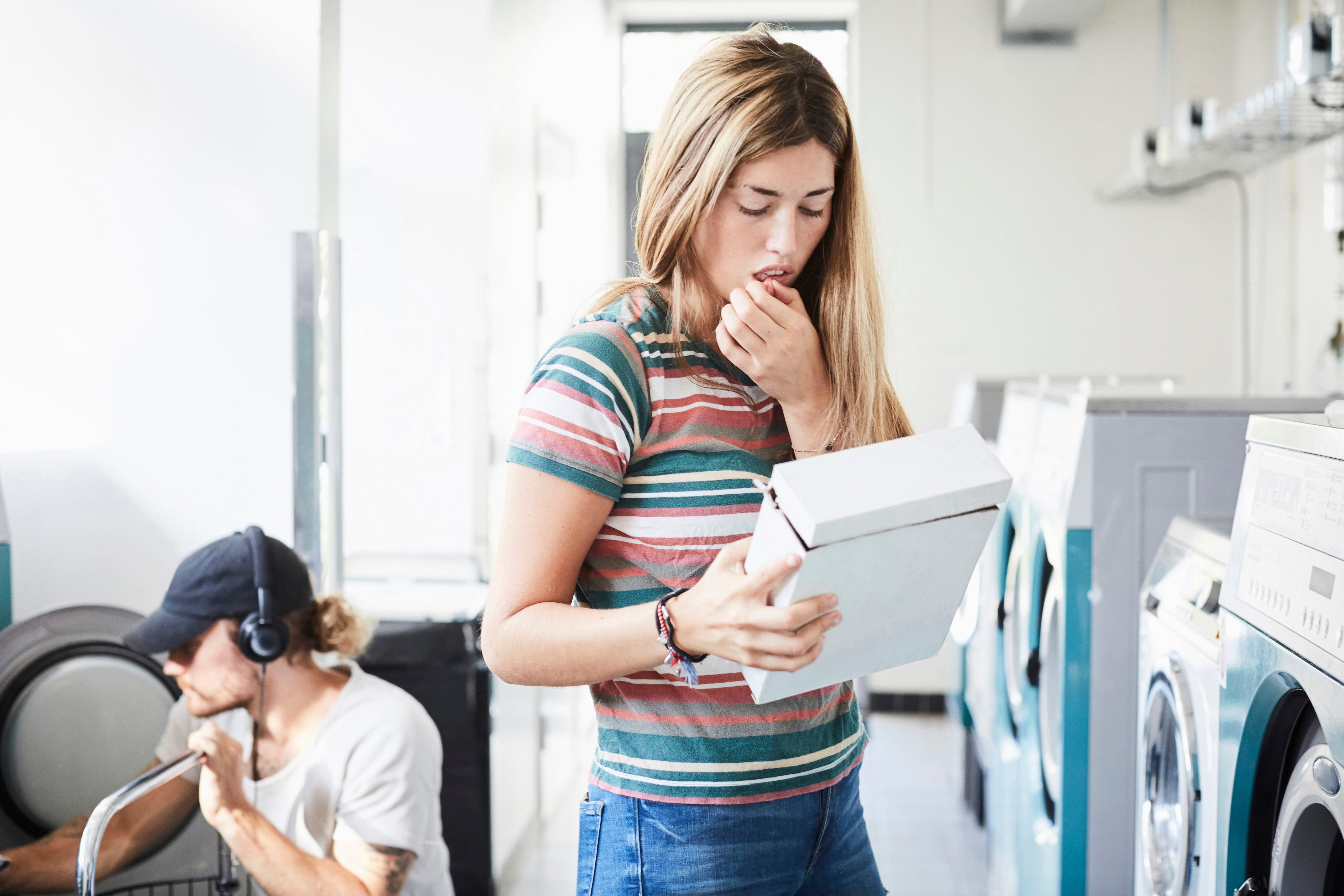 Woman in striped shirt looks at a box in a laundromat, appearing puzzled. In the background, a person tends to a washing machine