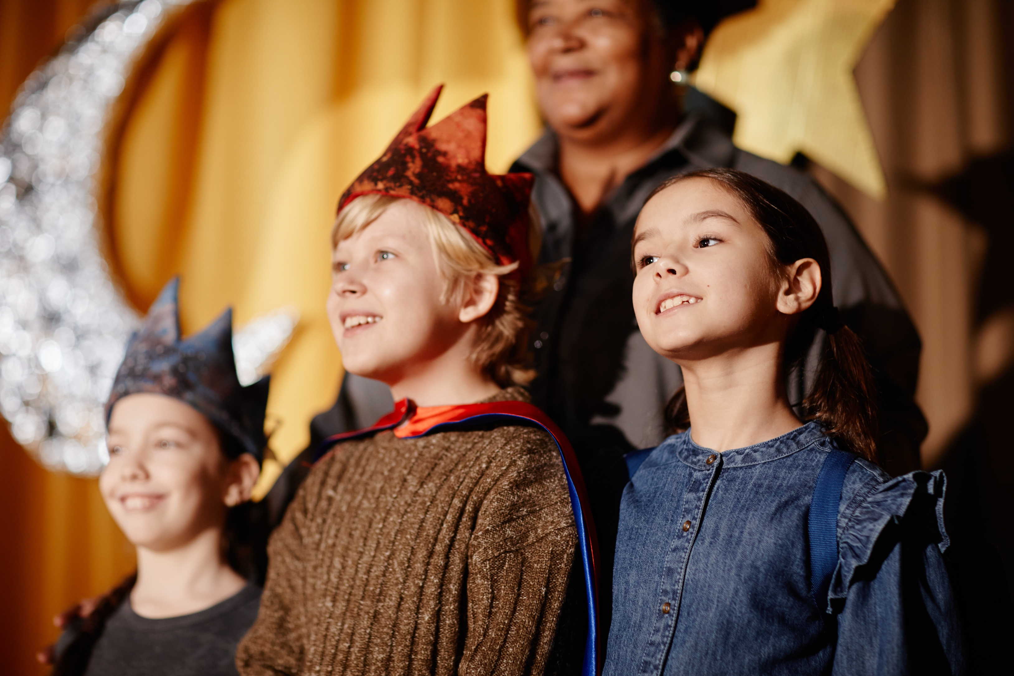 Children in costume wearing crowns, participating in a school play, stand on stage looking forward. Adults in background