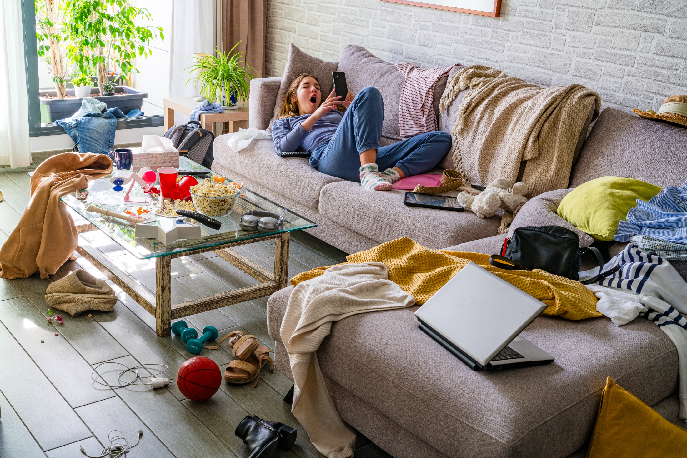 Person in casual clothing yawns on a cluttered couch, surrounded by scattered clothes, snacks, and electronics. The room is messy but cozy