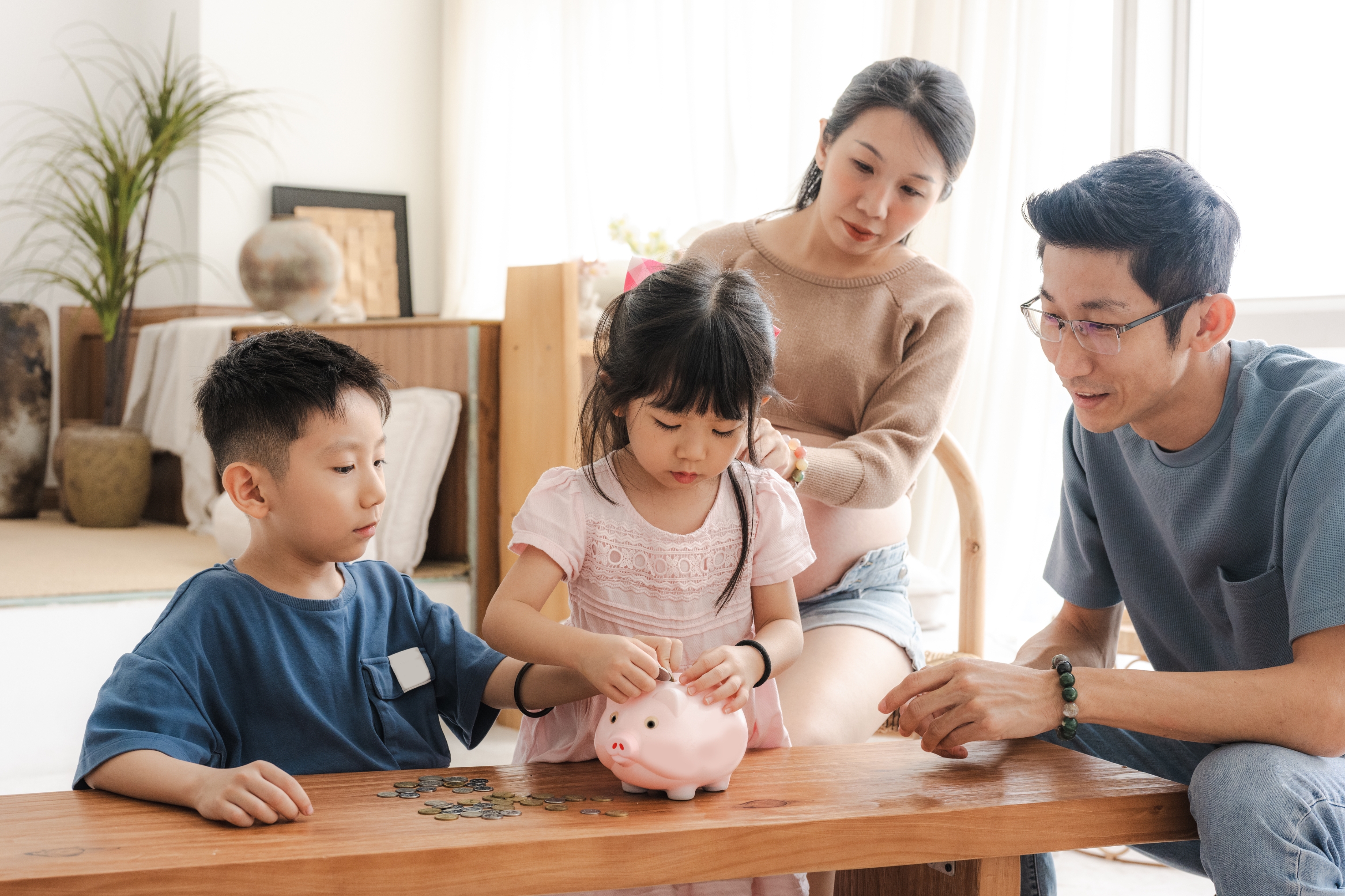 Family of four gathers around a wooden table as two children put coins into a pink piggy bank, indicating a family activity about saving money