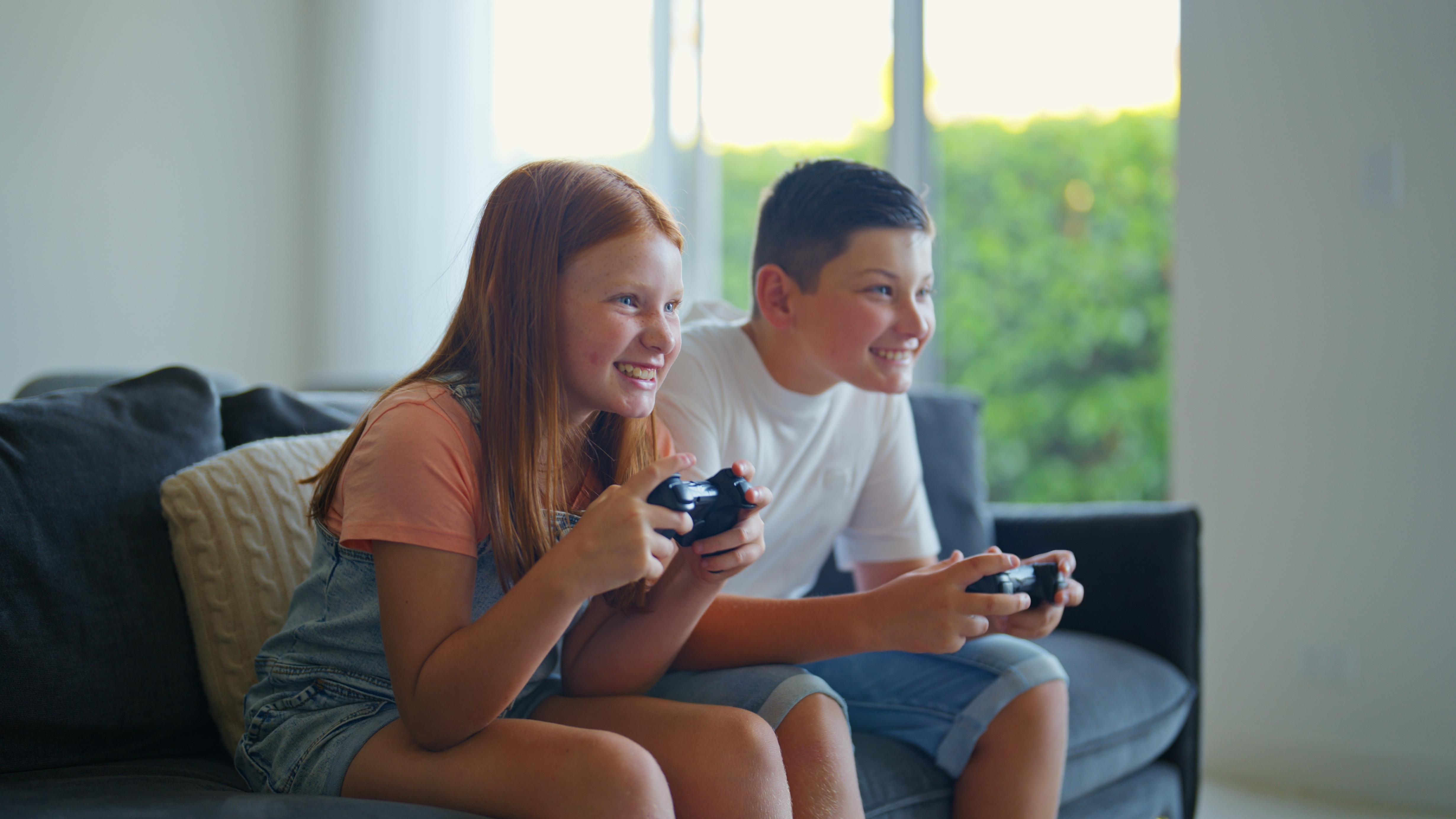 Two kids sitting on a couch, enthusiastically playing video games. They are focused and smiling, holding game controllers
