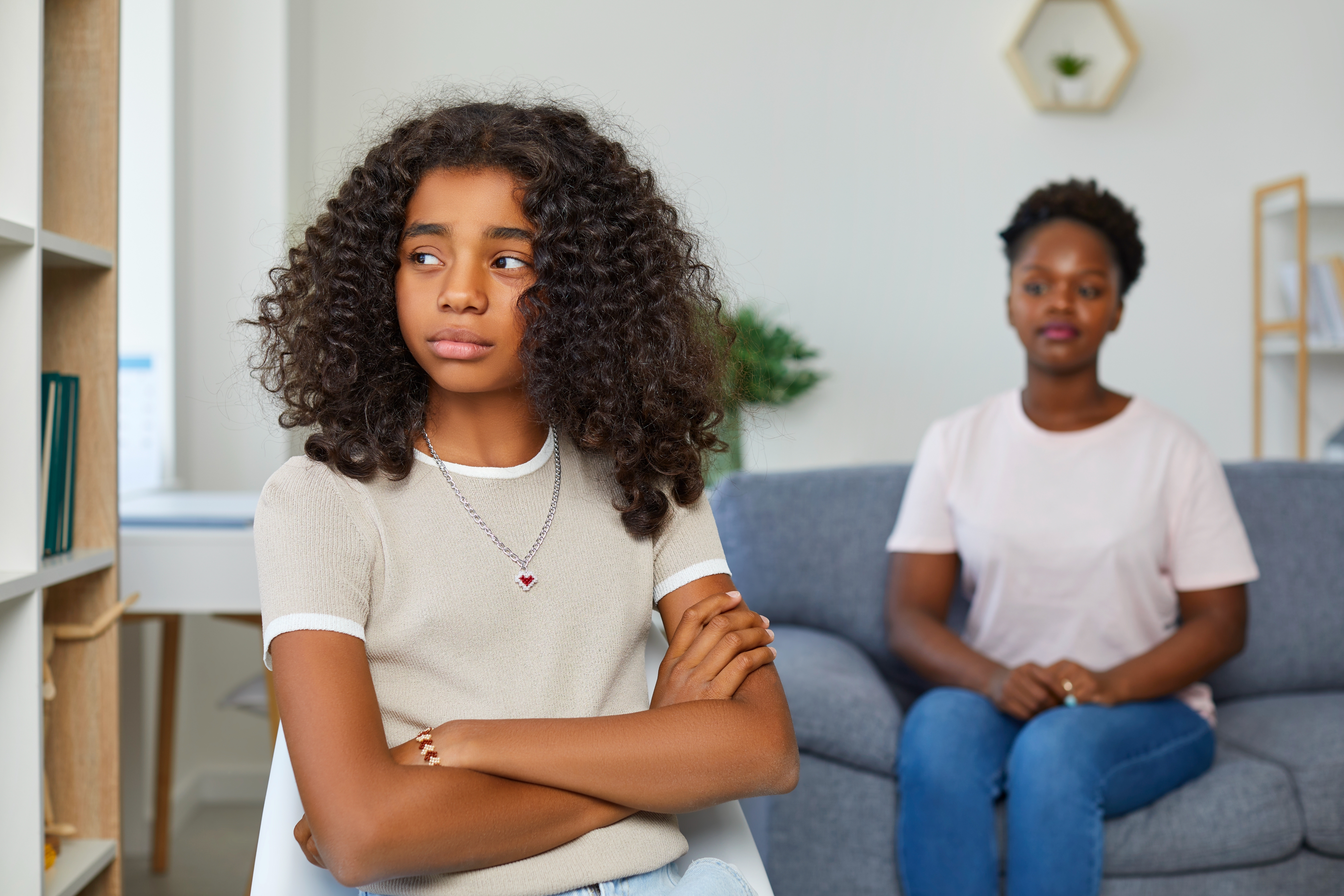 Teen girl with curly hair, arms crossed, looks away in thought; woman in background sits on couch, observing her quietly