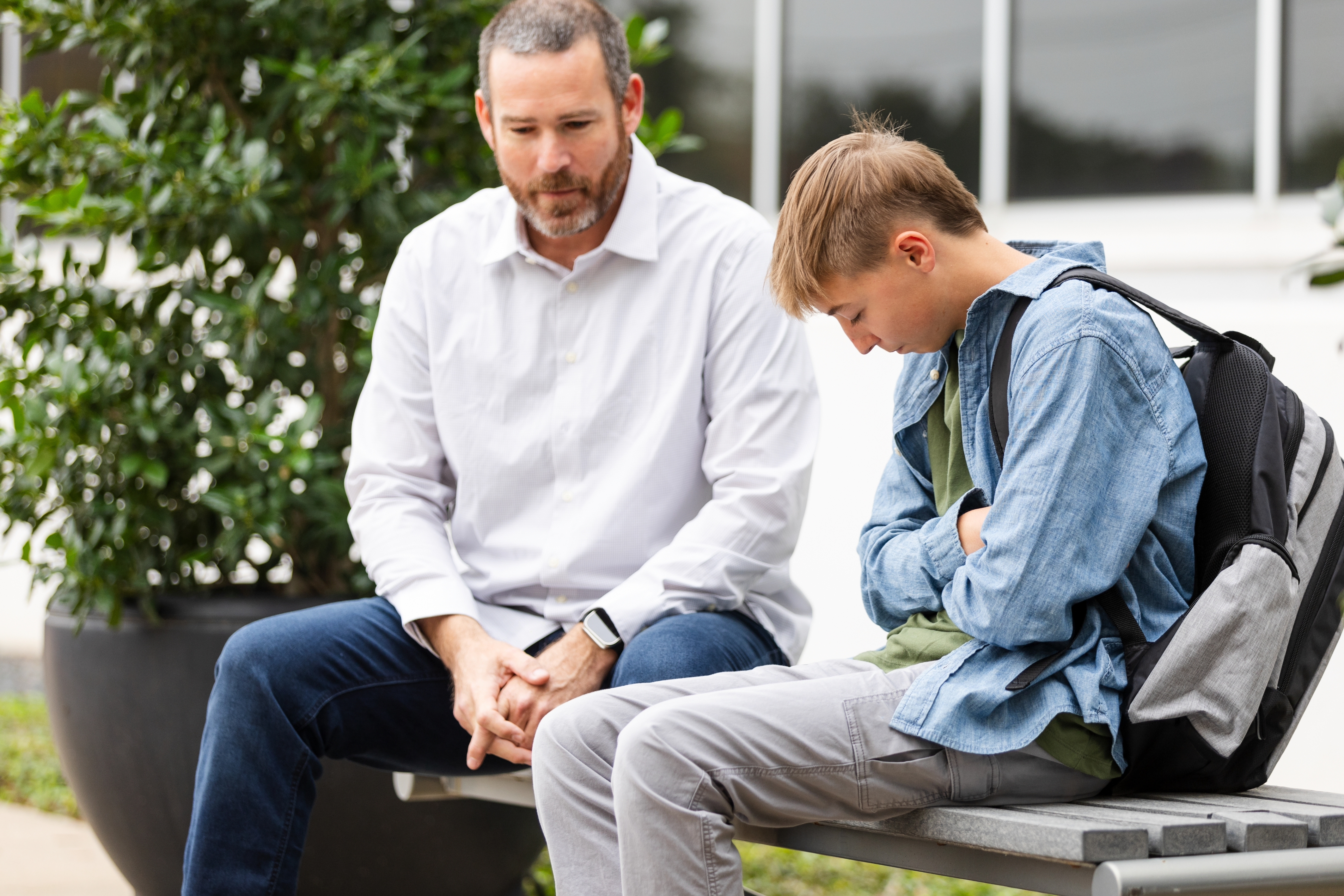 Man and teenager sitting on a bench, engaged in serious conversation. The man is attentive, while the teenager looks down, appearing thoughtful