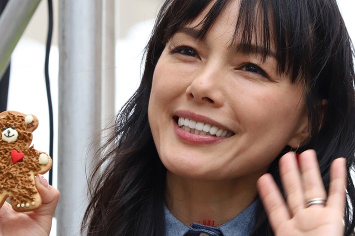 女性が笑顔でクマの形をしたクッキーを手に持ち、手を振っています。カジュアルな服装。