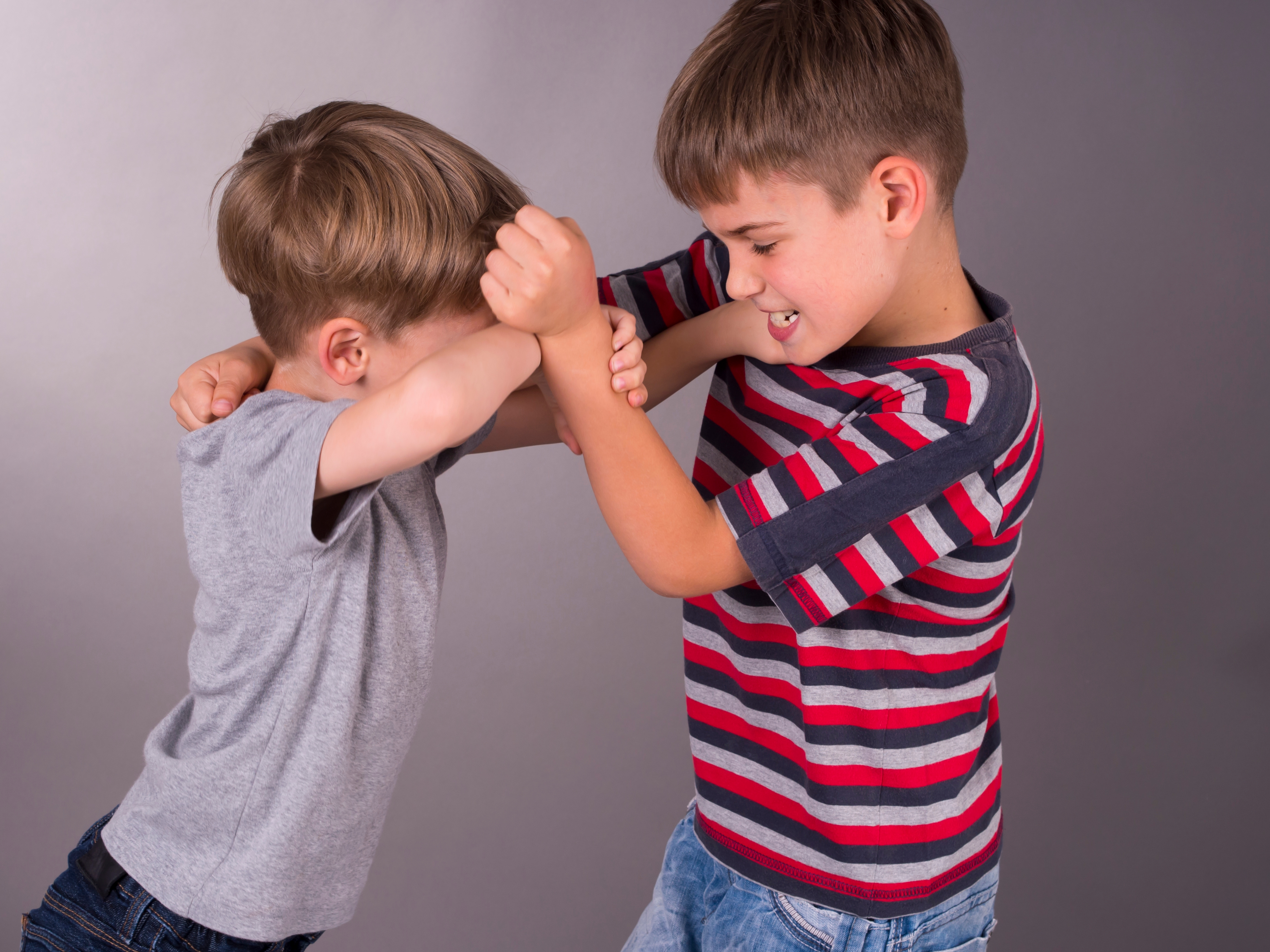 Two young boys are playfully wrestling indoors. One wears a striped shirt, and the other a plain one. Both are smiling and appear to be having fun