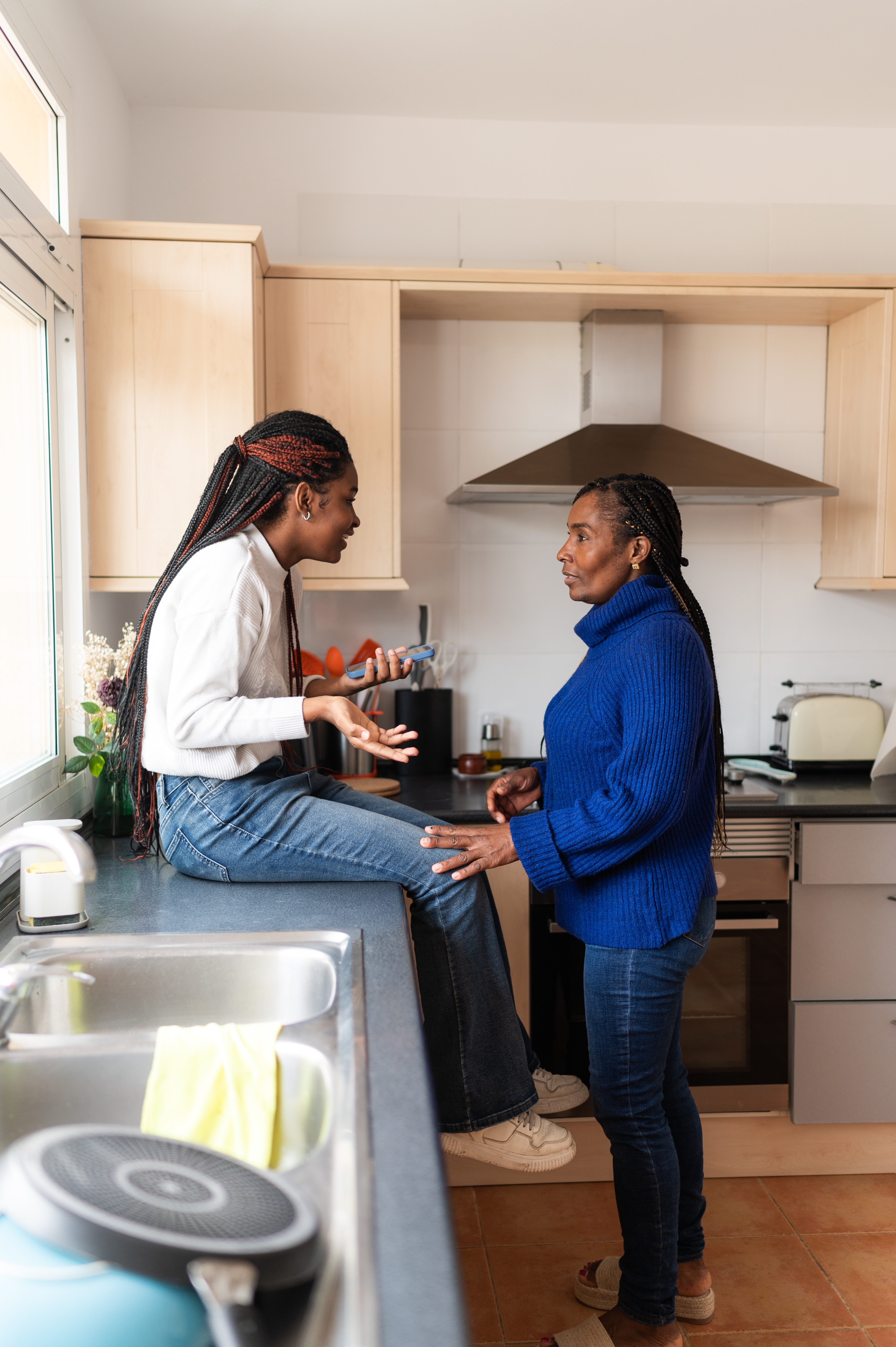 Two people talk in a kitchen; one sits on the counter, gesturing animatedly, while the other stands, listening attentively