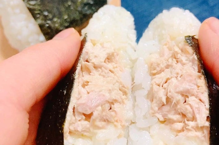 手で持ったツナ入りのおにぎりの断面が見える。背景にもう1つの海苔で包まれたおにぎりが置かれている。